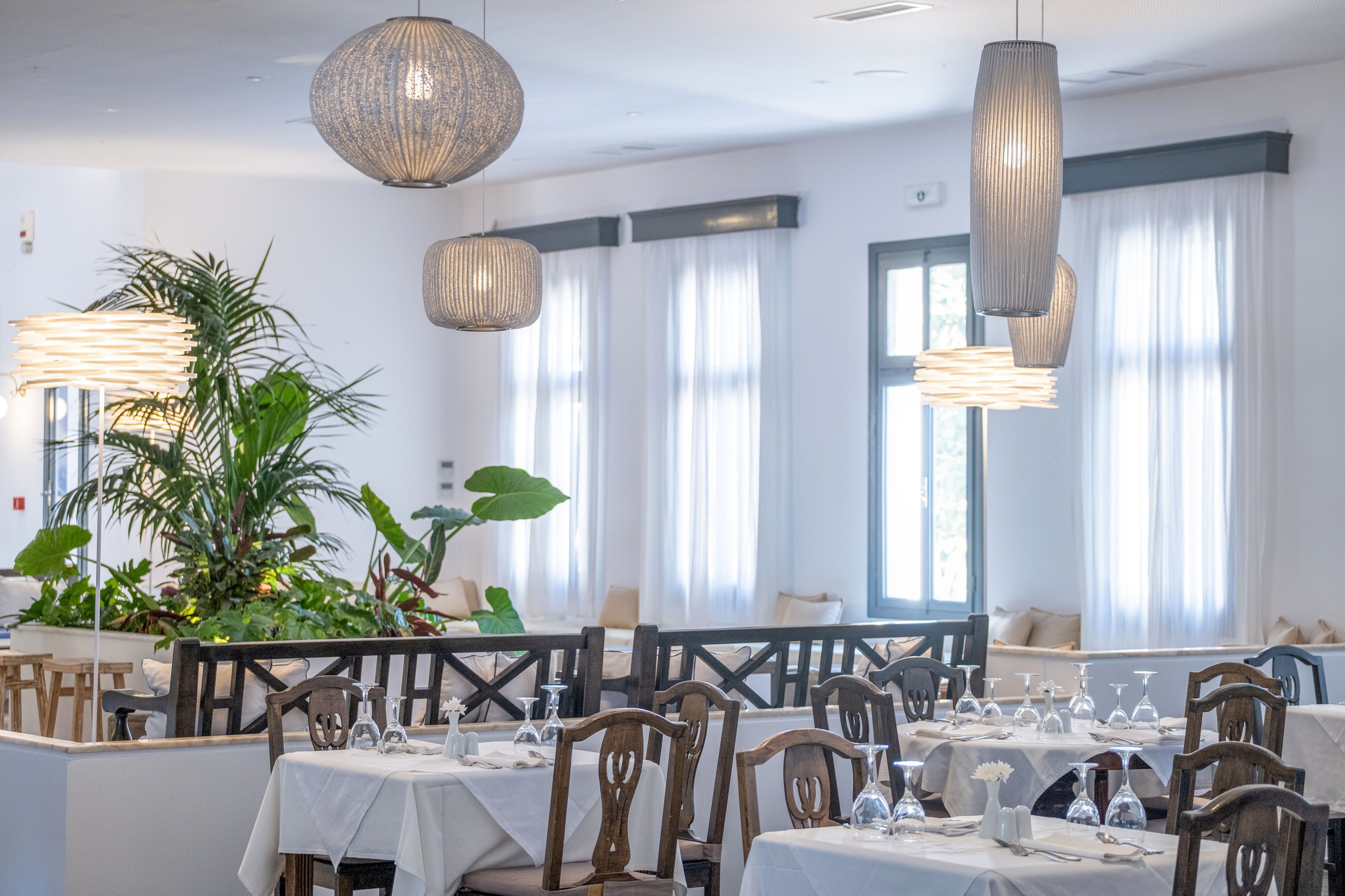 A contemporary restaurant dining room features neatly arranged tables covered with white tablecloths and set with glassware. Large windows with sheer white curtains allow soft natural light to fill the space. Decorative pendant lights hang from the ceiling, adding warmth and texture to the clean, minimalist design. Green indoor plants provide a fresh contrast to the predominantly white and neutral color palette.