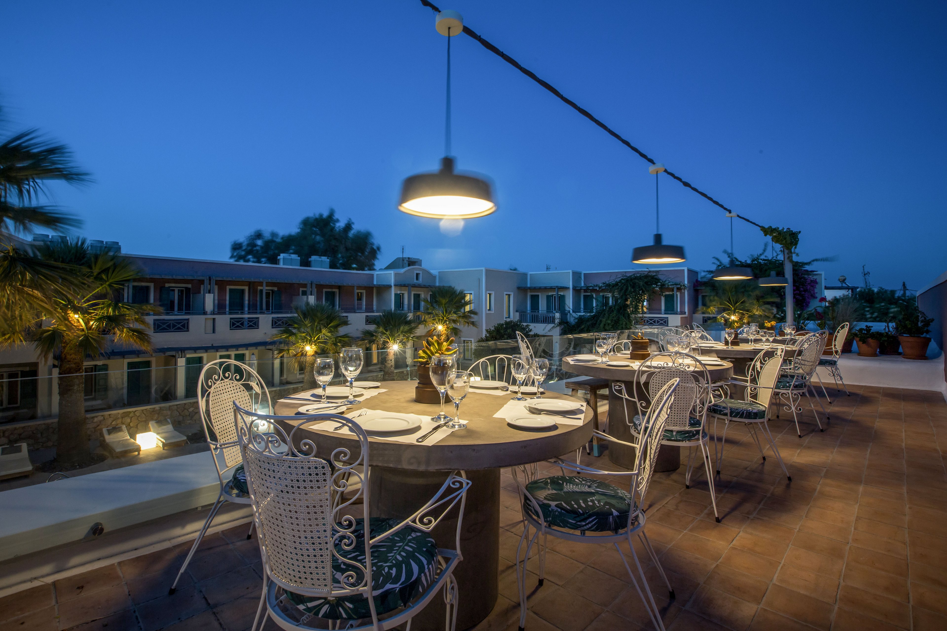An outdoor restaurant terrace is set for dinner on a rooftop at twilight. Round tables with white wrought-iron chairs are arranged along a tiled floor, each laid with plates, cutlery, glasses, and a small centerpiece. Warm pendant lights hang above the tables, contrasting with the deep blue evening sky. In the background, low-rise buildings and palm trees create a relaxed vacation atmosphere.