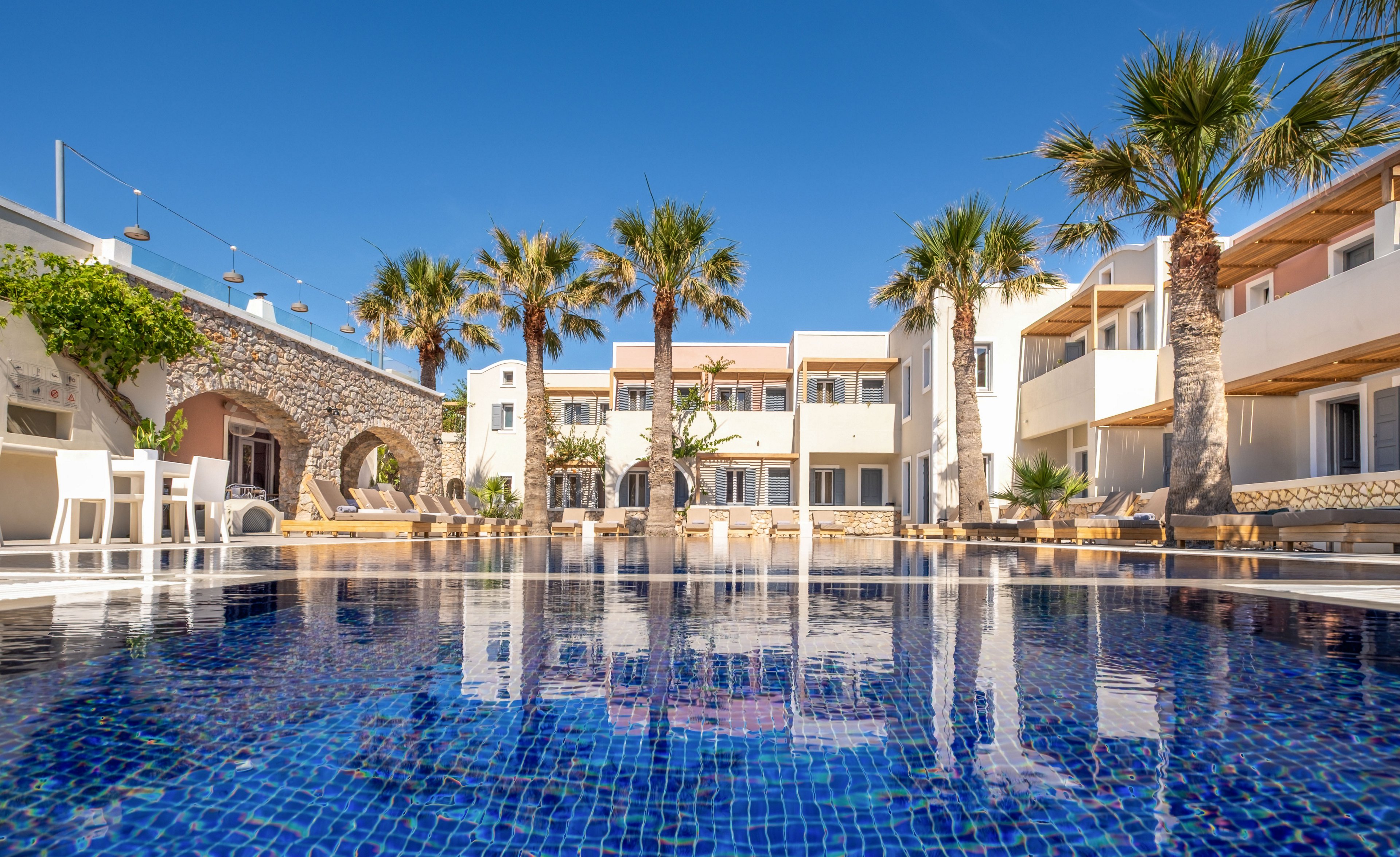 A tranquil resort courtyard features a clear blue swimming pool reflecting the surrounding buildings and palm trees. White and pastel-colored structures with balconies frame the pool, creating a relaxed vacation atmosphere. Lounge chairs and shaded seating areas line the poolside under a bright, cloudless sky. Stone archways and tiled surfaces add Mediterranean architectural charm to the scene.