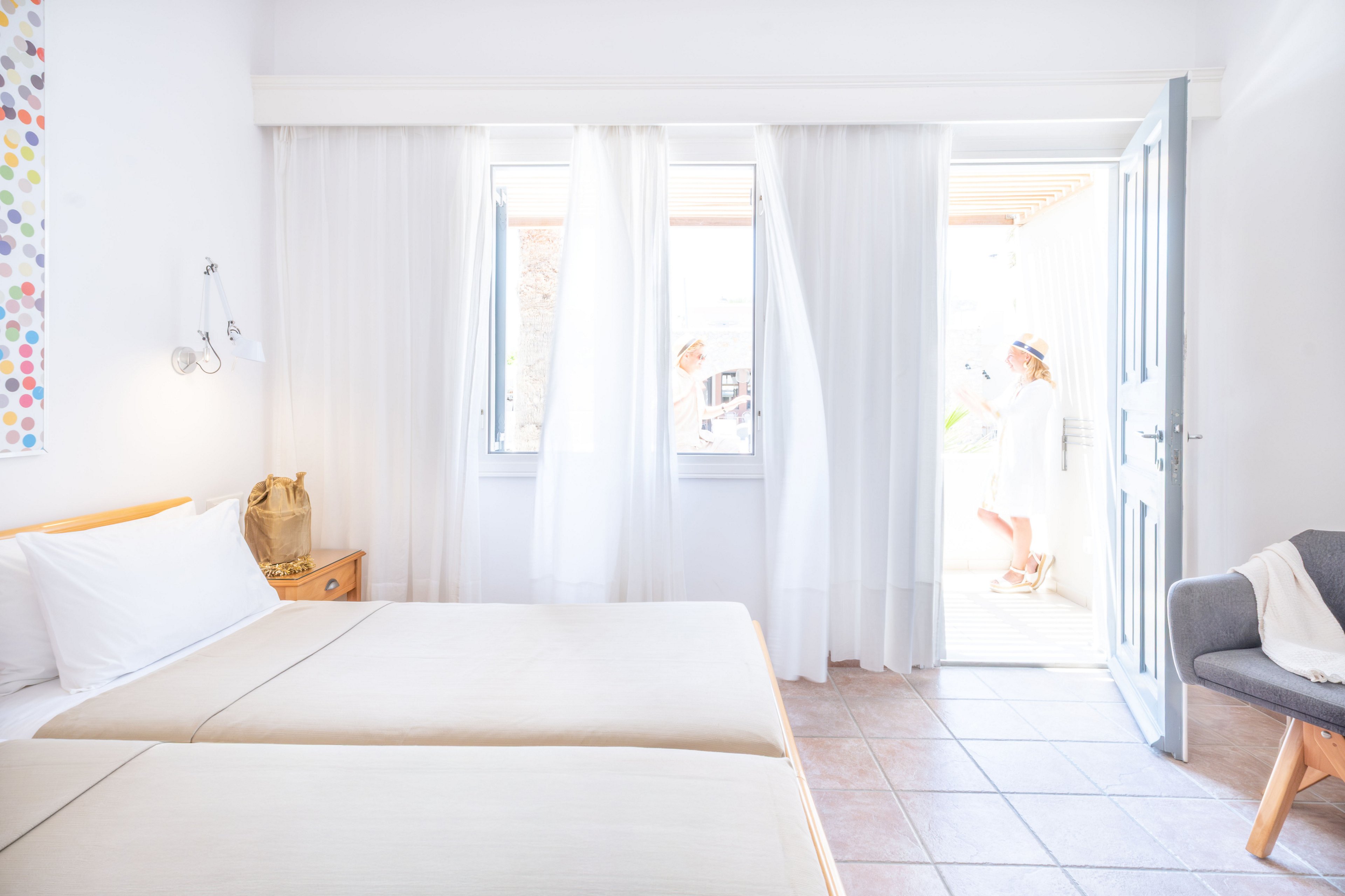 A sunlit hotel room features two neatly made twin beds against a white wall. Sheer white curtains hang in front of large windows, allowing soft daylight to fill the space. A small wooden nightstand with a woven bag and a gray armchair with a pillow add simple, modern decor. Through the open door, two people in light summer clothing are visible in the bright outdoor area, suggesting a relaxed vacation setting.