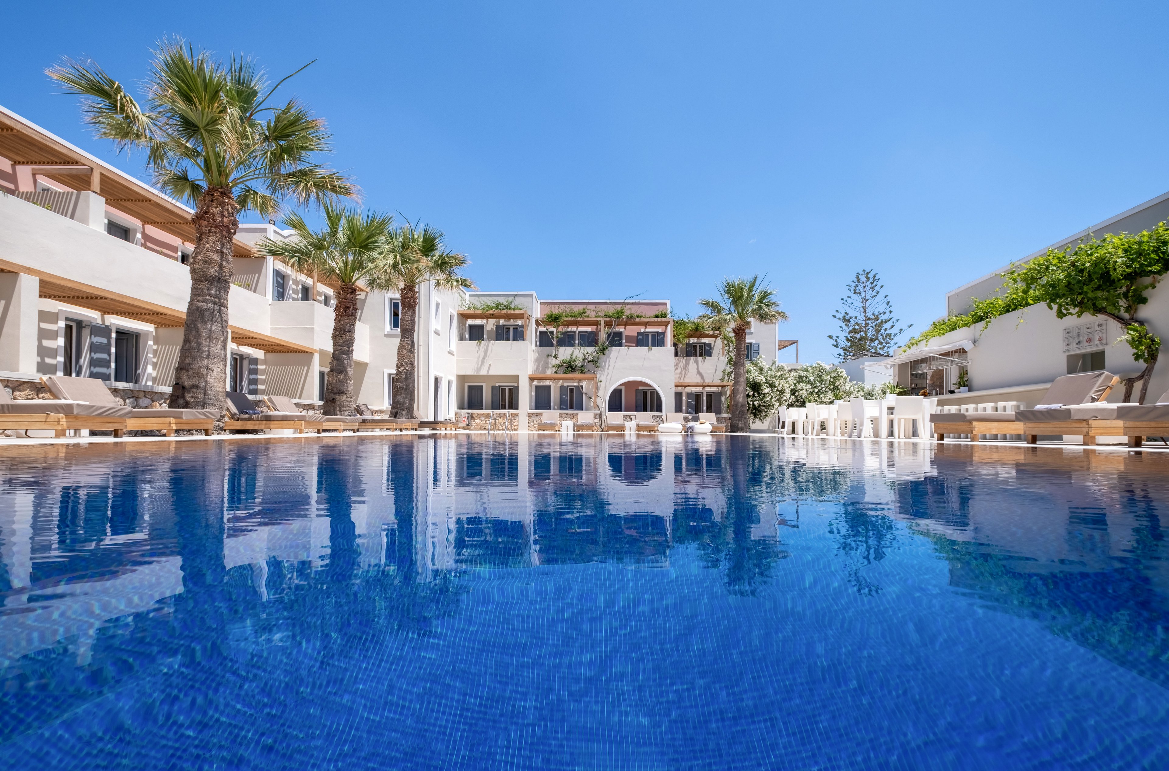 A large blue swimming pool stretches through the center of a modern resort courtyard. White and beige buildings with balconies and terraces surround the water on both sides. Palm trees and greenery add a tropical touch under a clear blue sky. Sun loungers line the poolside, suggesting a relaxing vacation setting. The calm water reflects the architecture and bright daylight.