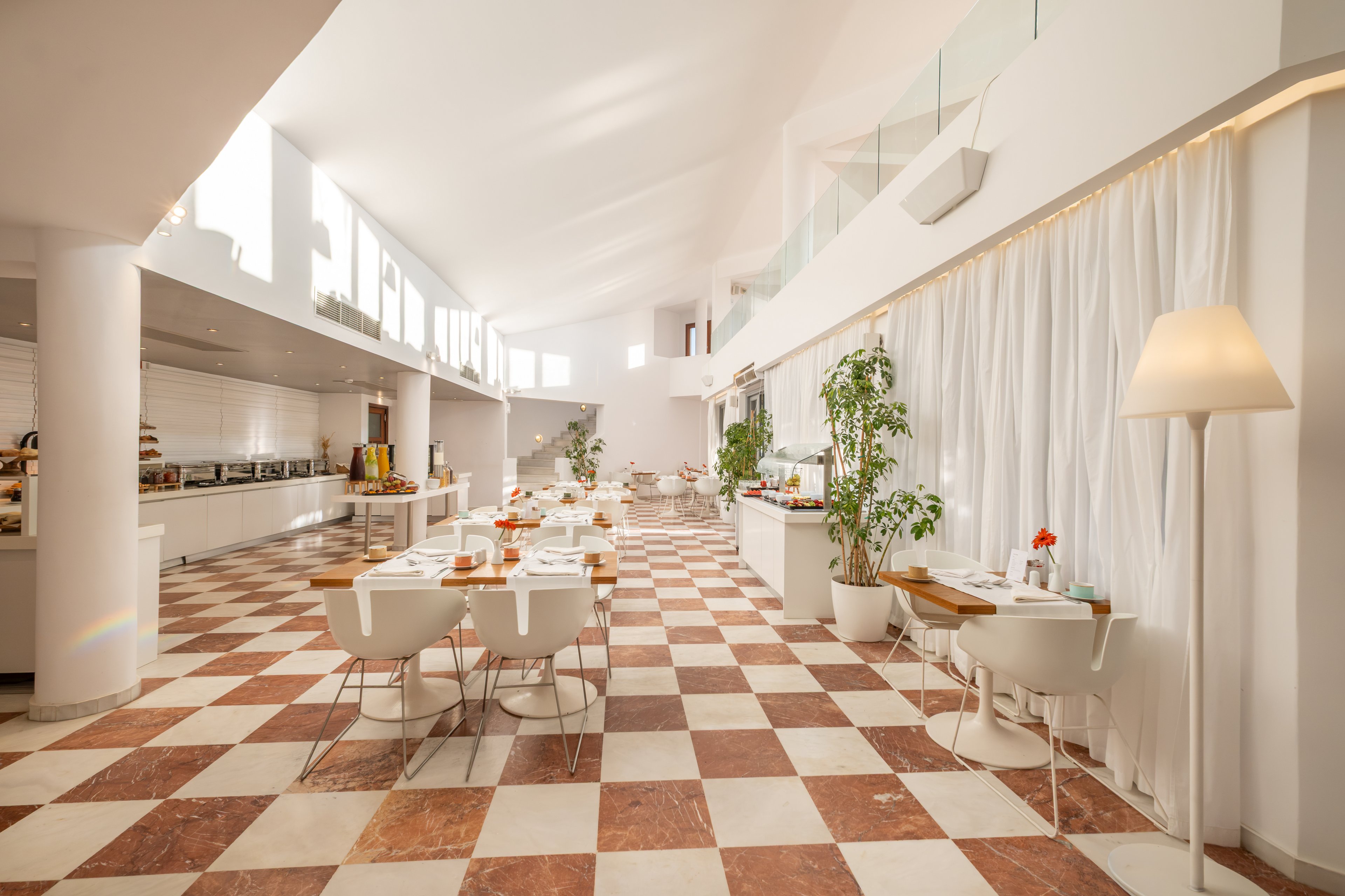 Spacious contemporary restaurant interior with a long buffet counter along one wall. Sunlight streams through high windows, illuminating checkered tile flooring and white furniture. Tables are set with plates, glasses, and small floral centerpieces, creating a welcoming dining atmosphere. Potted plants, soft curtains, and warm lighting add a relaxed, airy feel to the space.