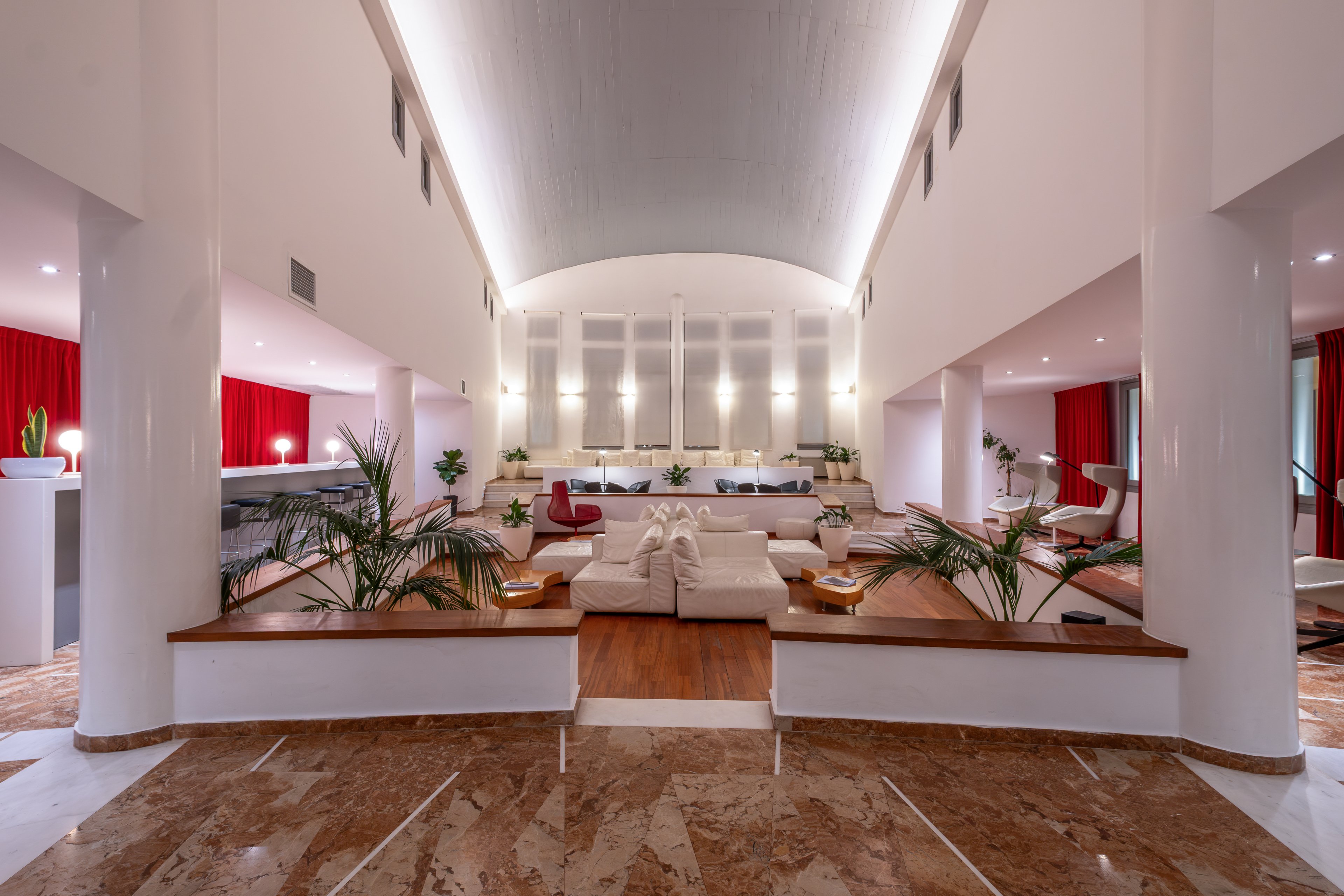 Spacious contemporary lobby interior features white sofas and armchairs arranged in a central lounge area. The setting includes tall white columns, polished stone flooring, and warm wood accents. Potted green plants, soft ambient lighting, and red curtains add contrast and visual interest. The overall atmosphere is elegant, welcoming, and designed for guest relaxation.