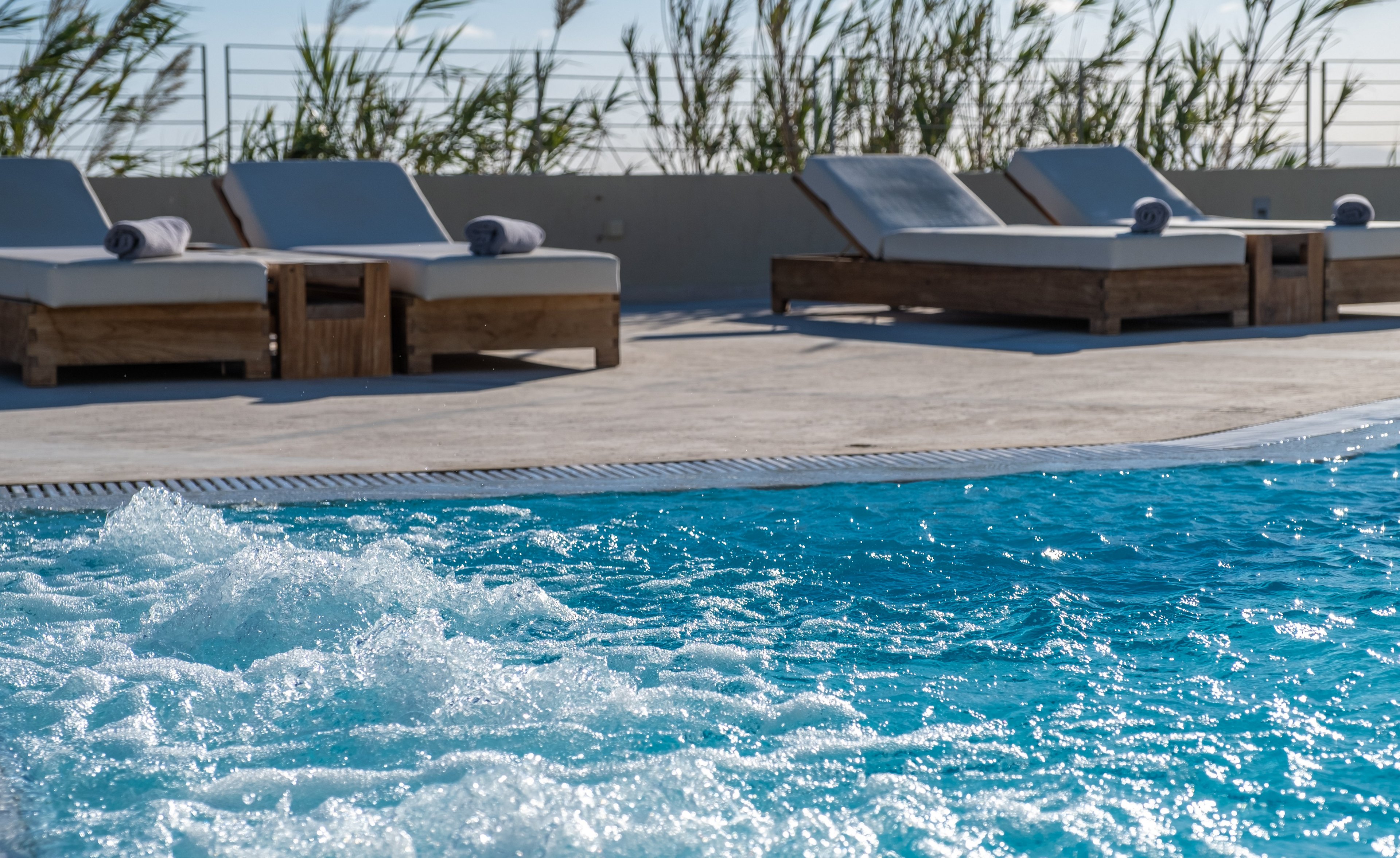 A clear blue swimming pool with bubbling water is seen in the foreground. Behind it, several cushioned wooden sun loungers line a light stone pool deck. Tall grasses and a simple railing frame the background, suggesting an upscale outdoor terrace. The scene evokes relaxation and resort-style leisure under bright daylight.