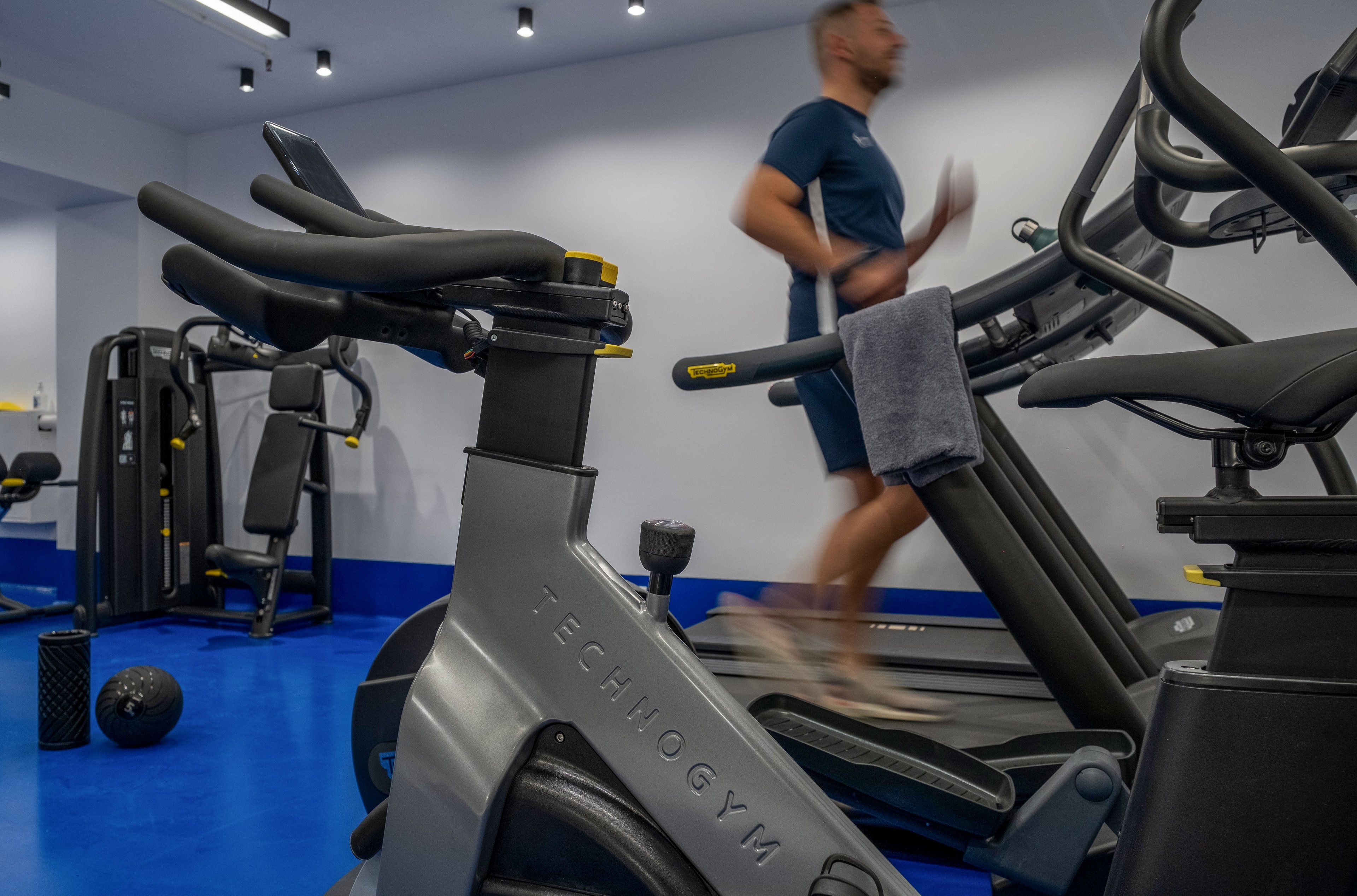 A man in athletic clothing runs on a Technogym treadmill in a modern indoor gym. The foreground shows a Technogym exercise bike with a towel draped over its handlebars. The gym features bright blue flooring, white walls, and ceiling spotlights, creating a clean and contemporary atmosphere. Additional fitness equipment, including a foam roller and weights, is visible in the background.
