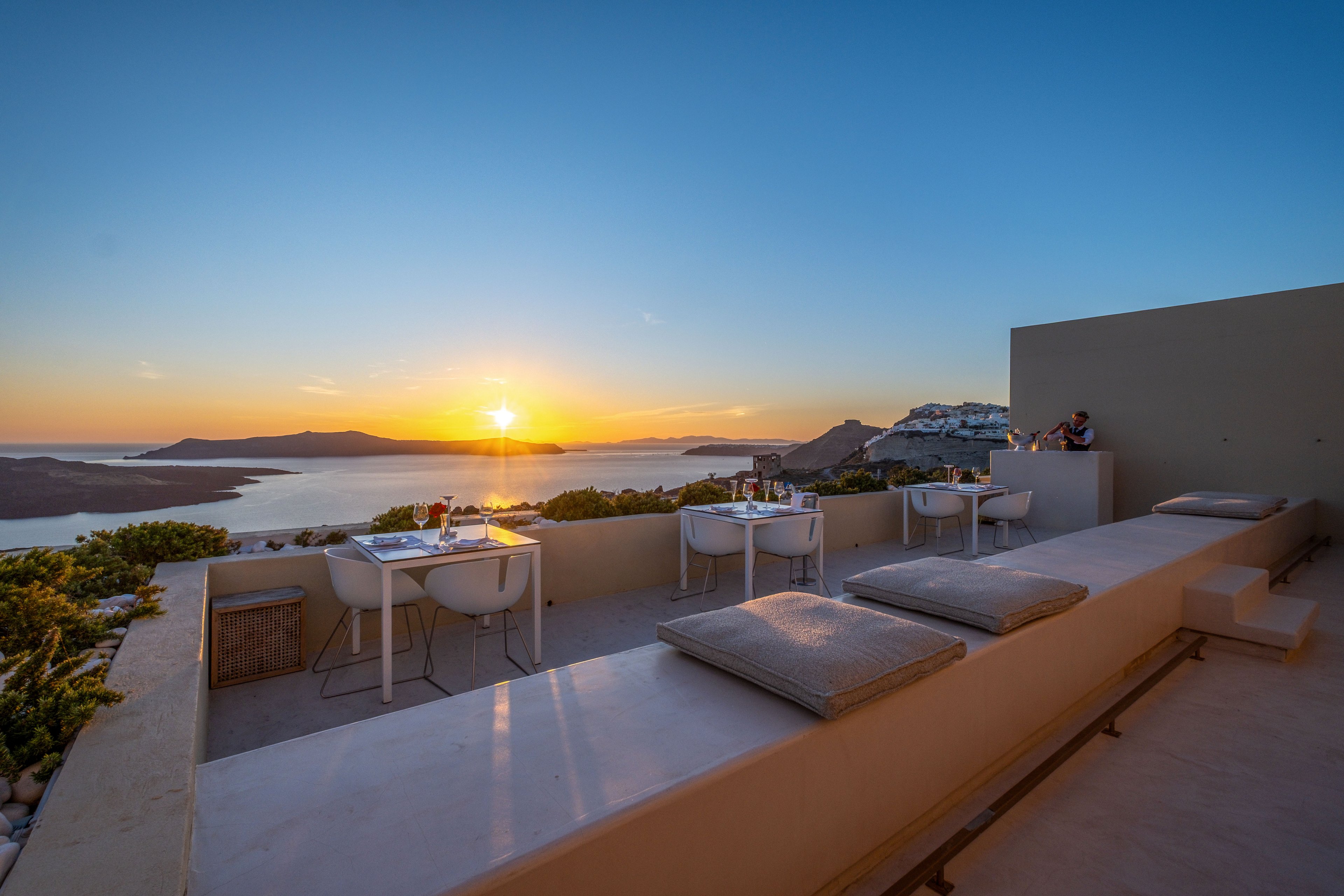 An elegant outdoor terrace features modern tables and chairs set for dining, facing a dramatic coastal landscape at sunset. Soft cushions line a long bench, inviting relaxation while guests enjoy the panoramic sea view. The sun is low on the horizon, casting warm golden light over the water and distant cliffs. The scene conveys a tranquil, romantic atmosphere ideal for evening leisure.