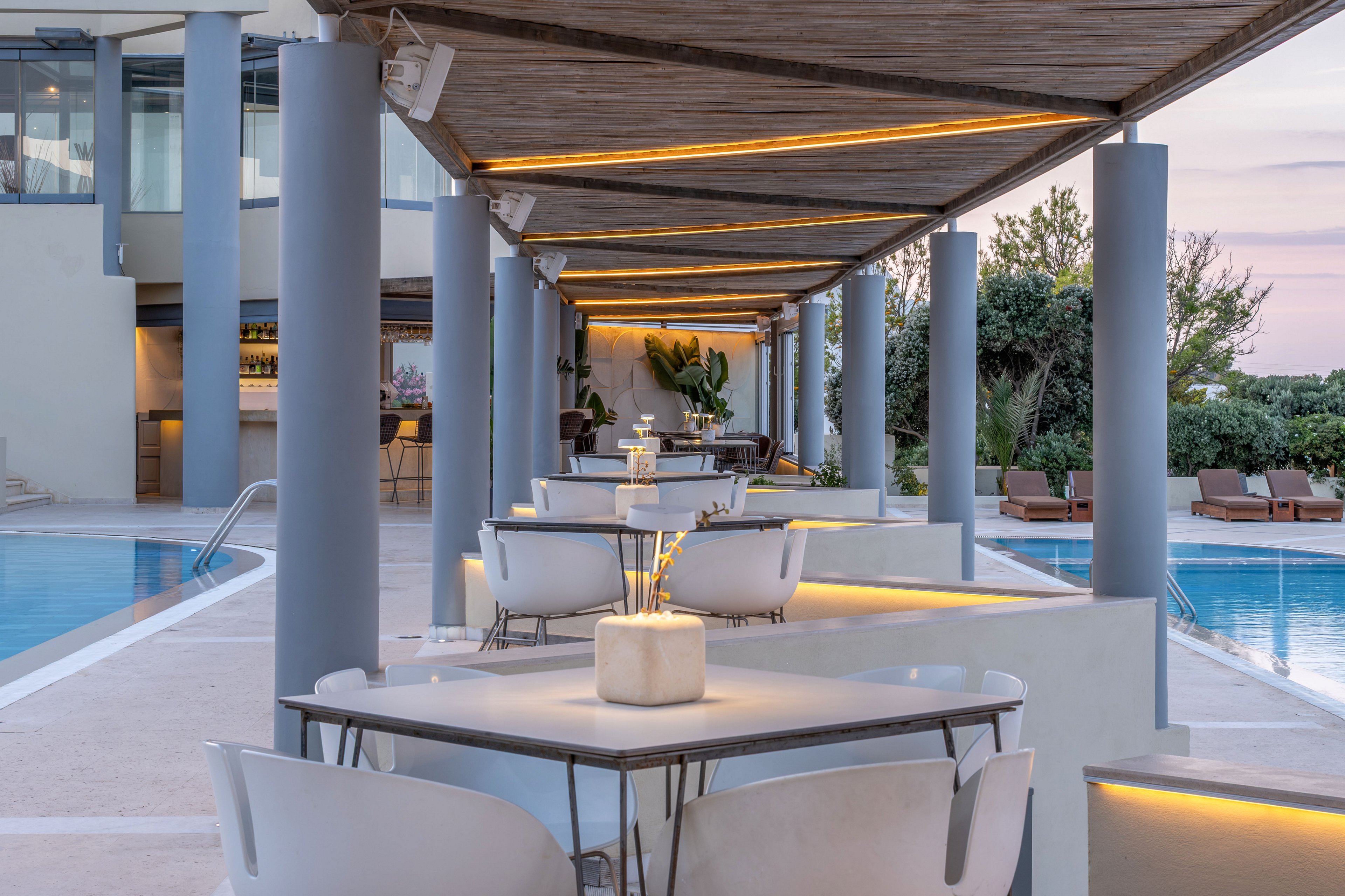 Contemporary outdoor dining tables line a covered terrace beside a clear blue swimming pool. Sleek white chairs and minimalist table settings create a refined, resort-style atmosphere. Warm linear lights under the wooden canopy contrast with the cool tones of the concrete columns and water. In the background, a bar area and sun loungers suggest a relaxed vacation setting surrounded by greenery.