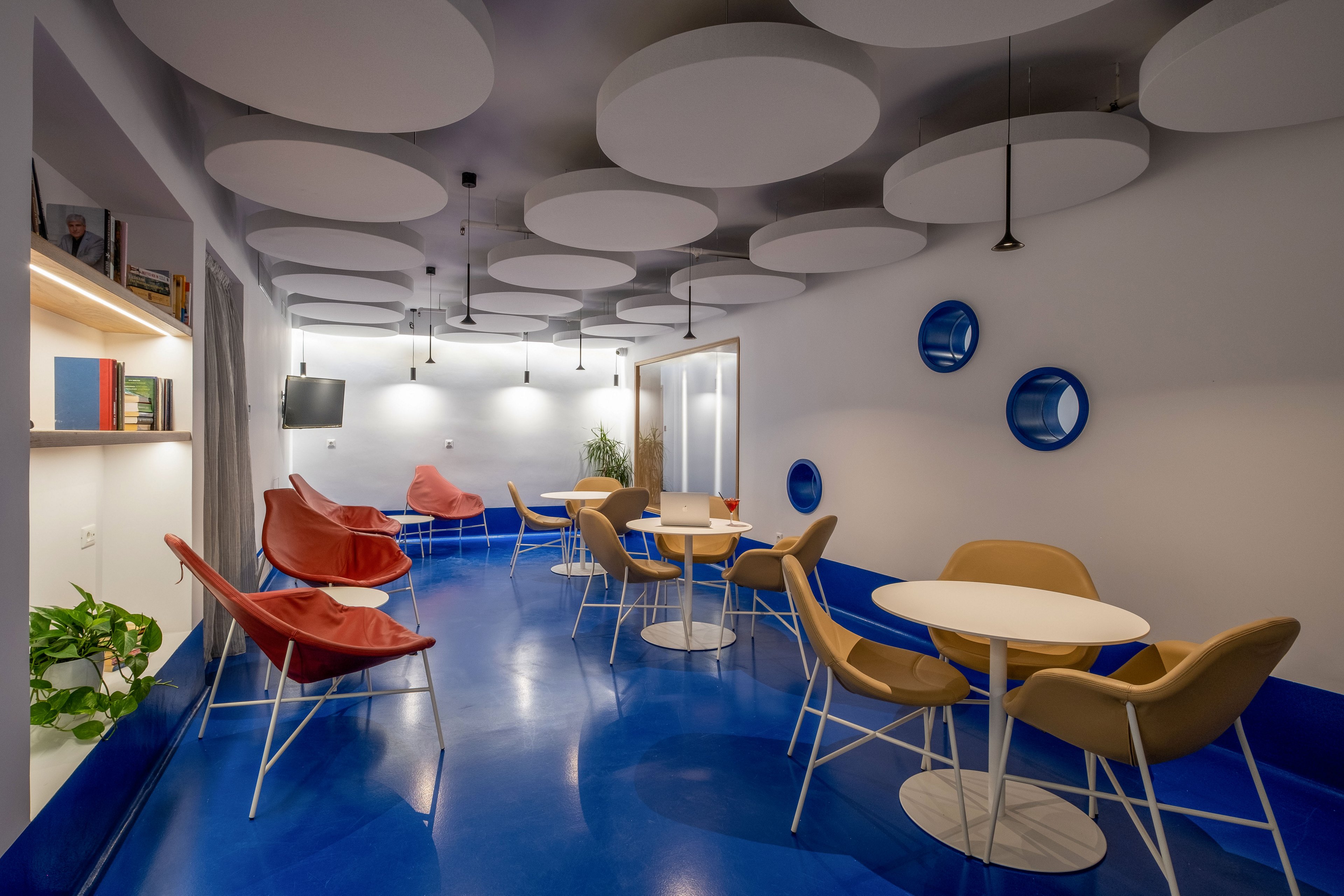 Contemporary lounge space features round tables with tan and red chairs on a glossy blue floor. Circular white ceiling panels and hanging pendant lights create a distinctive modern look. A wall-mounted television, bookshelves, and decorative round wall accents add visual interest. The room appears calm and inviting, suitable for informal meetings or relaxation.