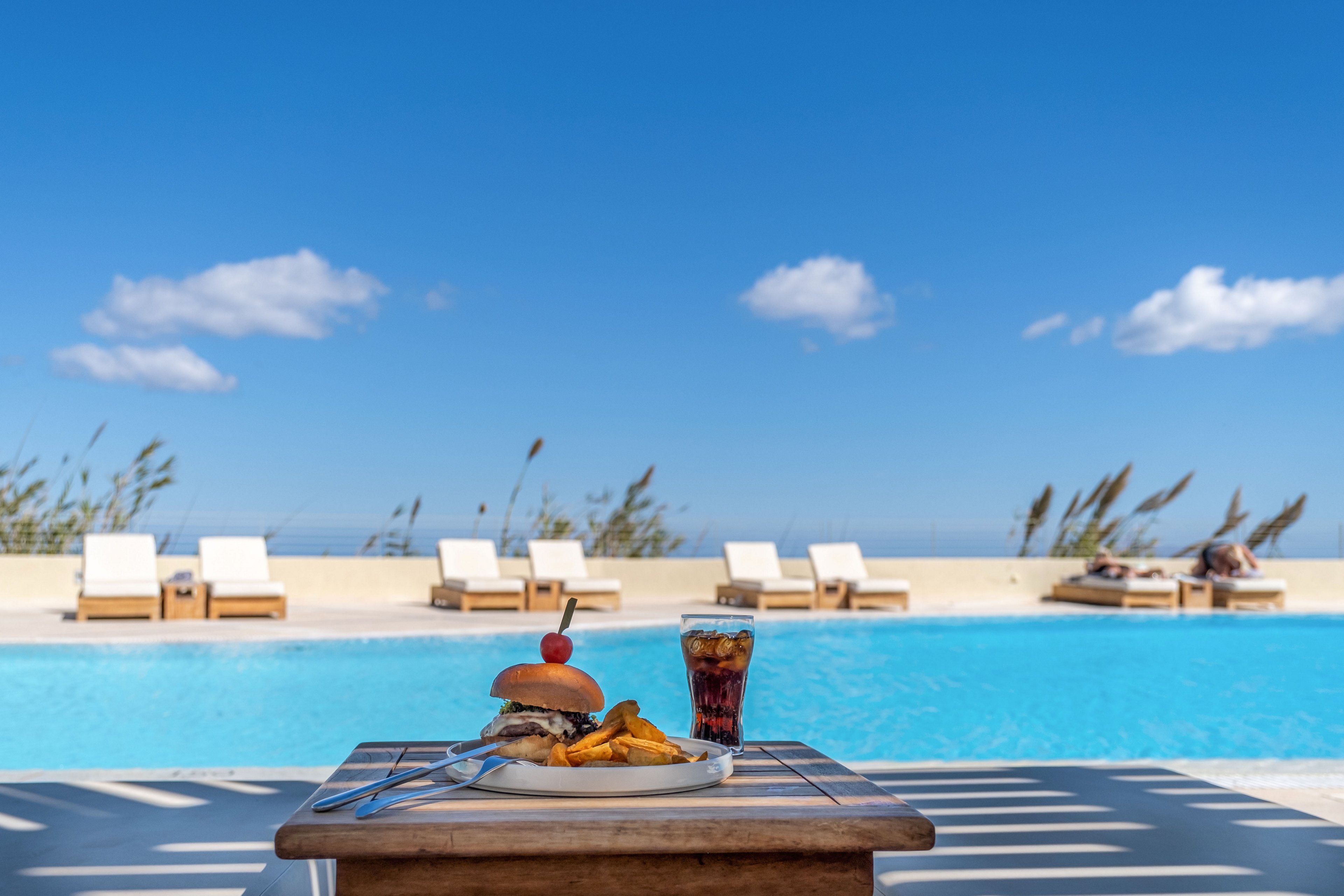 A plate with a burger and fries is set on a wooden table beside a clear blue swimming pool. A glass of soda stands next to the meal, suggesting a relaxed vacation lunch. In the background, white lounge chairs line the pool deck under a bright blue sky with scattered clouds. The scene evokes a sunny, tranquil resort atmosphere by the water.