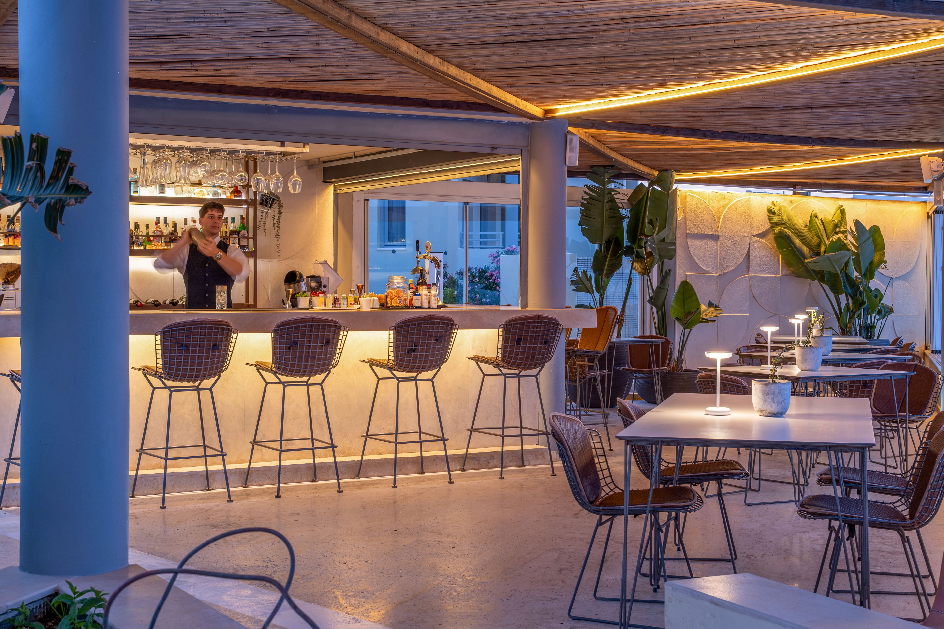 A bartender stands behind a sleek illuminated bar mixing drinks in an open-air lounge. Contemporary tables and woven bar stools fill the foreground, each lit by small table lamps that create a cozy evening ambiance. Warm linear lighting accents the wooden ceiling while large potted plants soften the modern architecture. The setting suggests a stylish, relaxed nightlife scene in a sophisticated urban or resort environment.