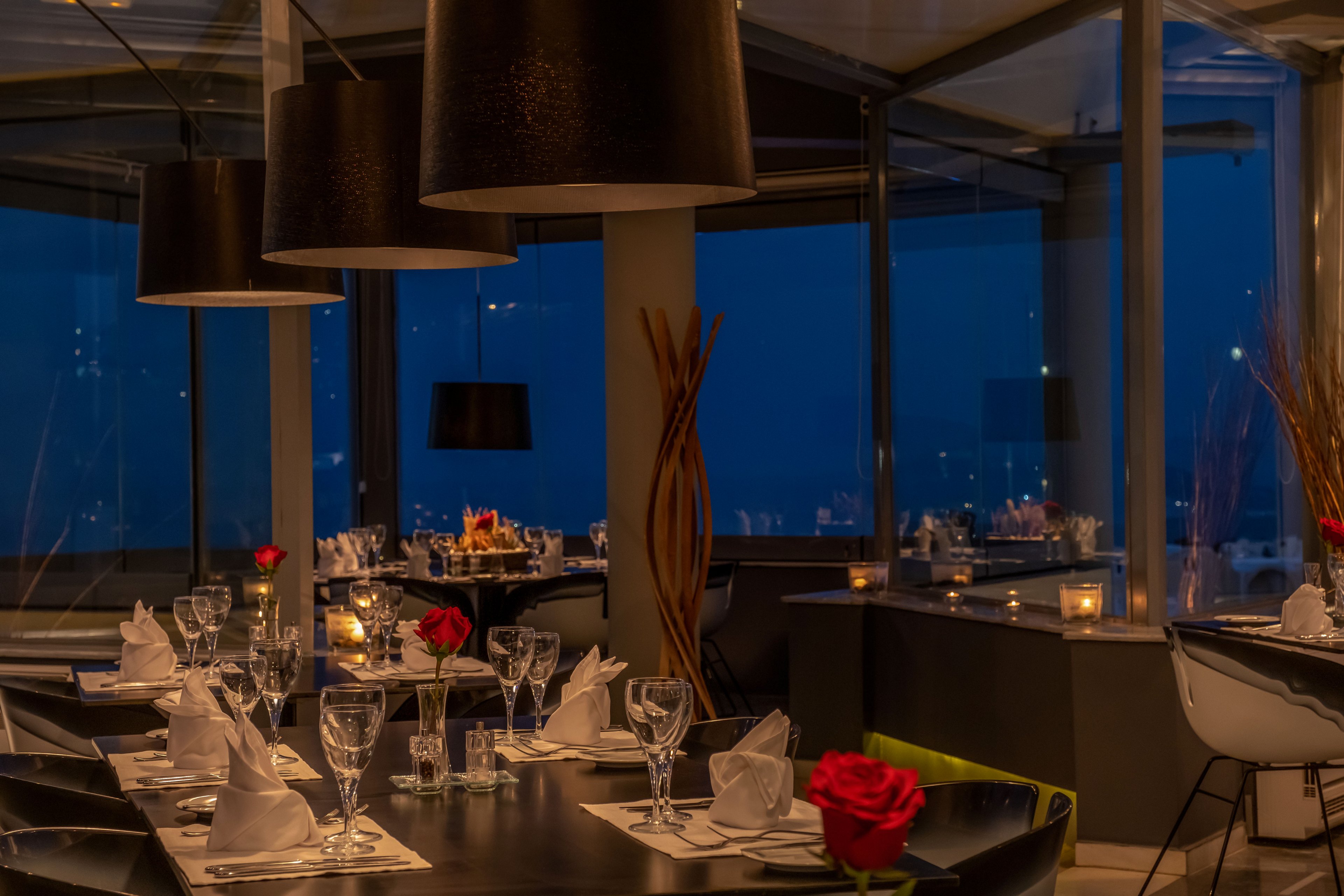 Elegant restaurant tables are set with folded white napkins, wine glasses, and single red roses in vases. Warm pendant lights and candles create a cozy, intimate atmosphere against large windows overlooking the night view. The dark tabletops contrast with the soft glow of the interior lighting. Decorative elements and reflections add depth and sophistication to the modern dining space.