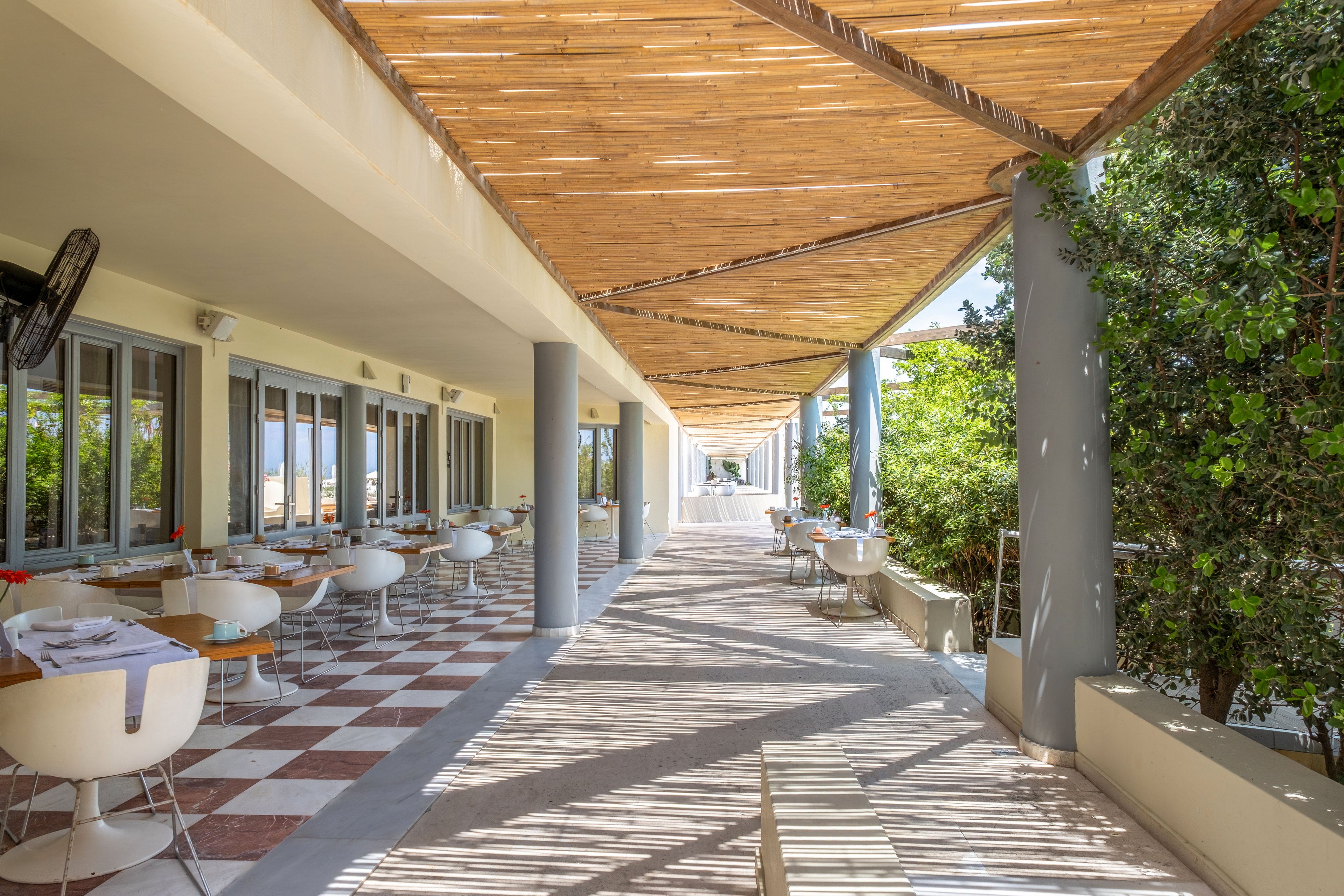 An open-air restaurant terrace features rows of modern white chairs and wooden tables set along a long corridor. A wooden pergola casts striped shadows on the tiled floor, creating a bright and inviting atmosphere. Large windows line one side of the space, while lush green foliage borders the other. The setting appears calm and ready for guests, with neatly arranged tableware and a clear view down the sunlit walkway.
