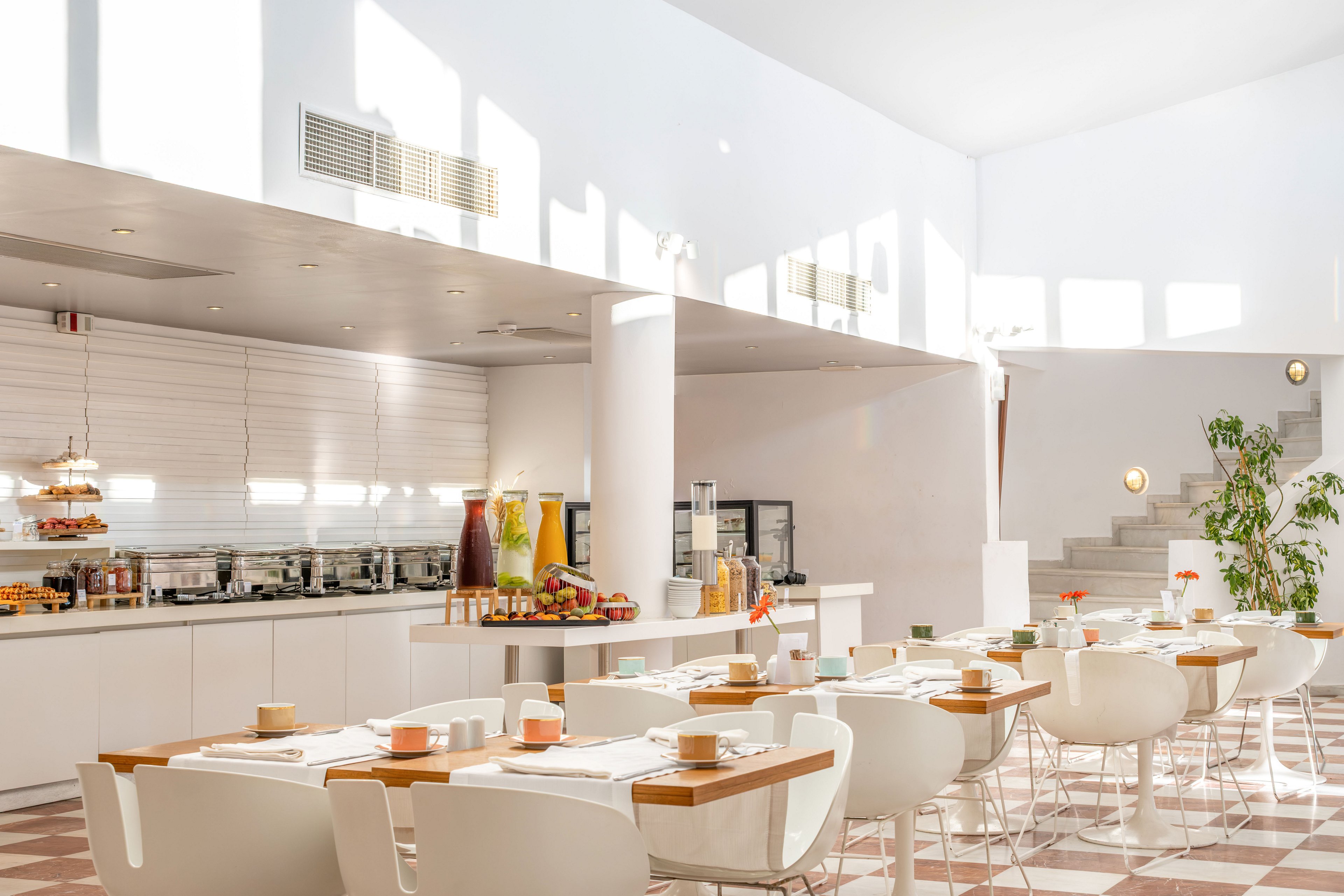 A sunlit cafeteria features neatly arranged white tables and chairs with place settings ready for guests. In the background, a breakfast buffet offers juices, pastries, and other food items on a long counter. Large windows and white walls create an airy, minimalist atmosphere, complemented by warm wooden table accents. The overall scene suggests a clean, contemporary dining space prepared for a morning meal.