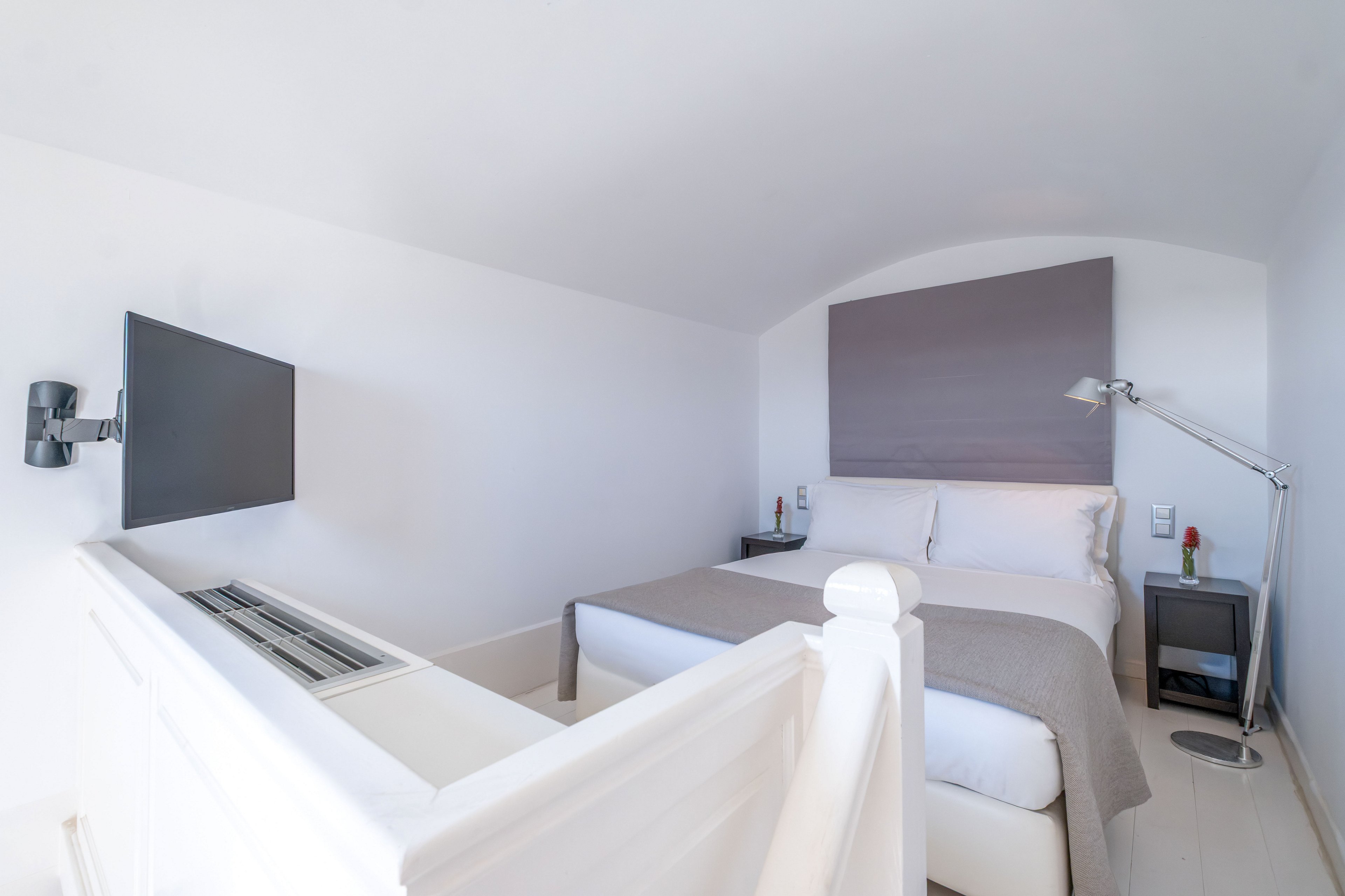 A compact modern bedroom features a neatly made double bed with white linens and a light gray throw. The room is predominantly white, with a soft curved ceiling and a large gray headboard panel above the bed. A wall-mounted flat-screen TV faces the bed near a white stair railing, and a sleek metal floor lamp stands beside a small bedside table. The overall design is clean, bright, and minimalist, emphasizing simplicity and comfort.