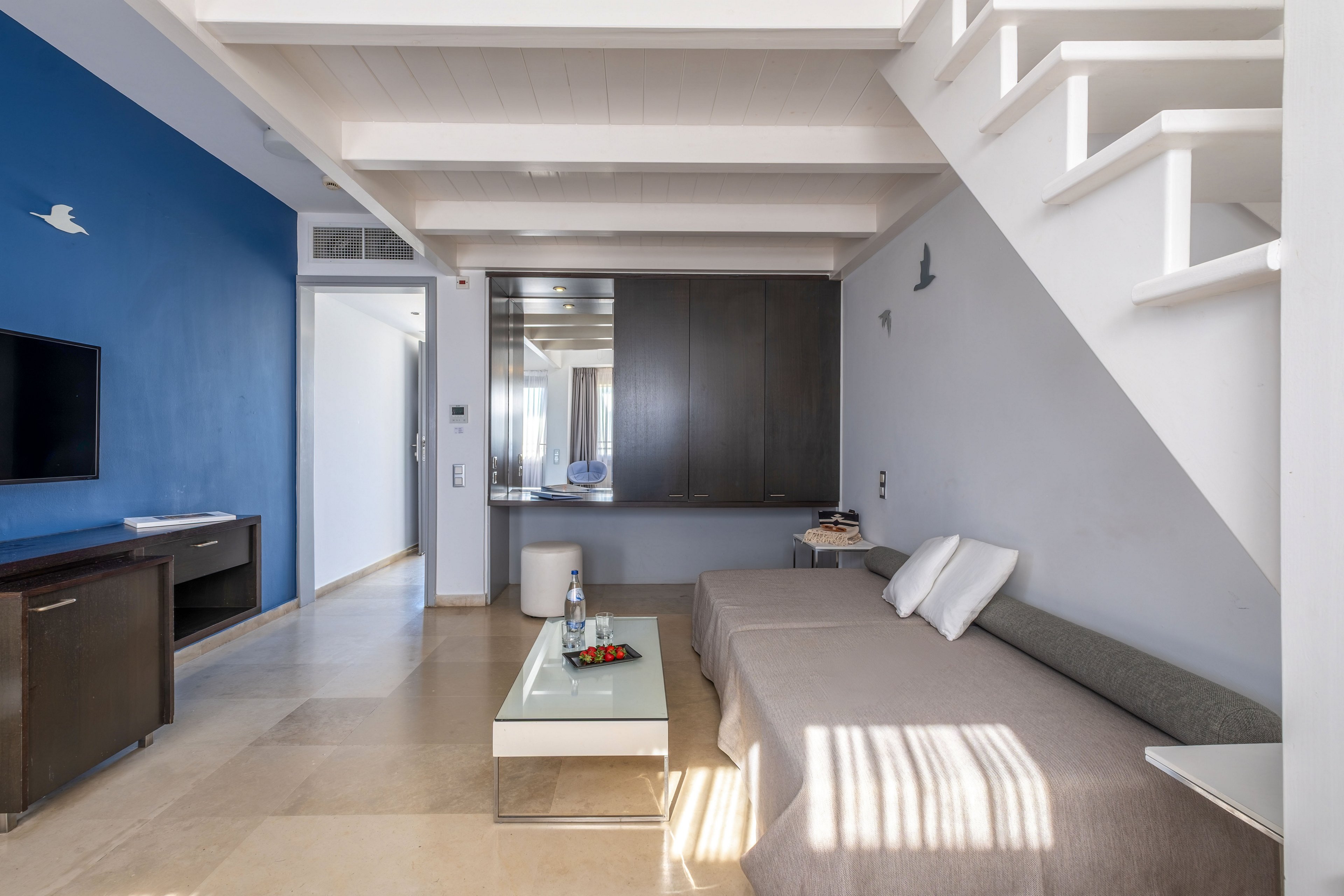 Contemporary living room interior featuring a low gray sofa and white cushions beneath a white staircase. A sleek glass coffee table with fruit and a drink sits on a light tiled floor. The space includes a blue accent wall with a mounted television and dark wood cabinetry. Sunlight streams through a window, casting shadows and creating a clean, airy atmosphere.
