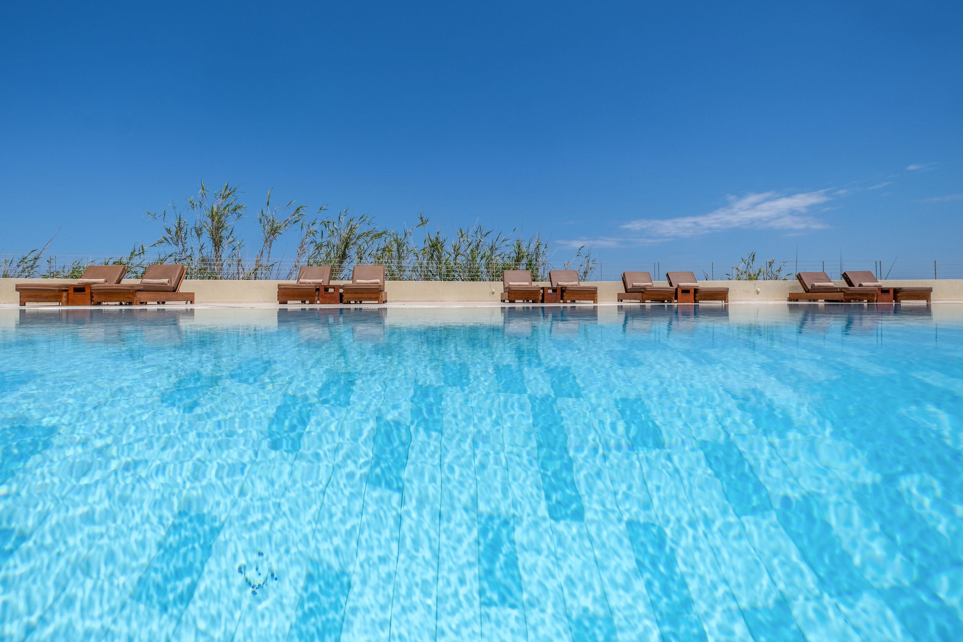A clear blue swimming pool stretches across the foreground under a bright cloudless sky. Wooden sun loungers with cushions line the far edge of the pool, suggesting a relaxing resort or hotel setting. Tall grasses or plants rise behind the loungers, adding a natural touch to the minimalist scene. The overall atmosphere is calm, inviting, and perfect for a summer vacation.