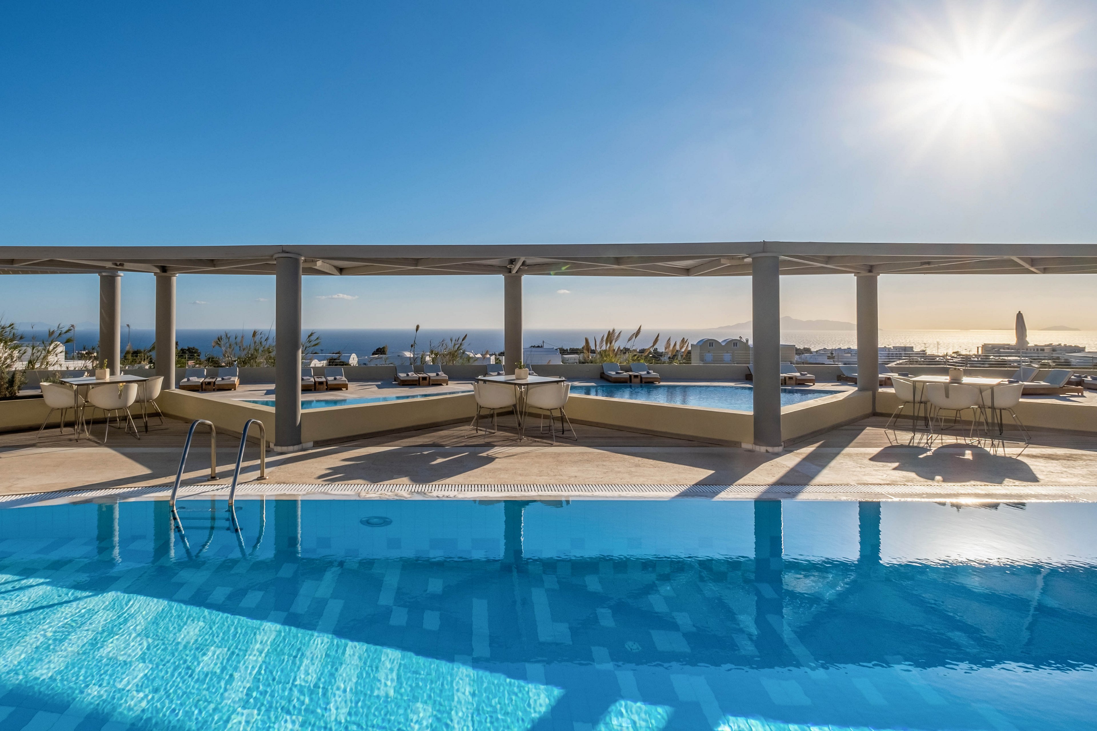 A serene infinity pool reflects the clear blue sky at a coastal resort. Lounge chairs and dining tables are arranged under a modern pergola structure, facing the ocean. The sun shines brightly near the horizon, casting long shadows across the terrace. Calm sea views and minimalist architecture create a tranquil, luxurious vacation atmosphere.