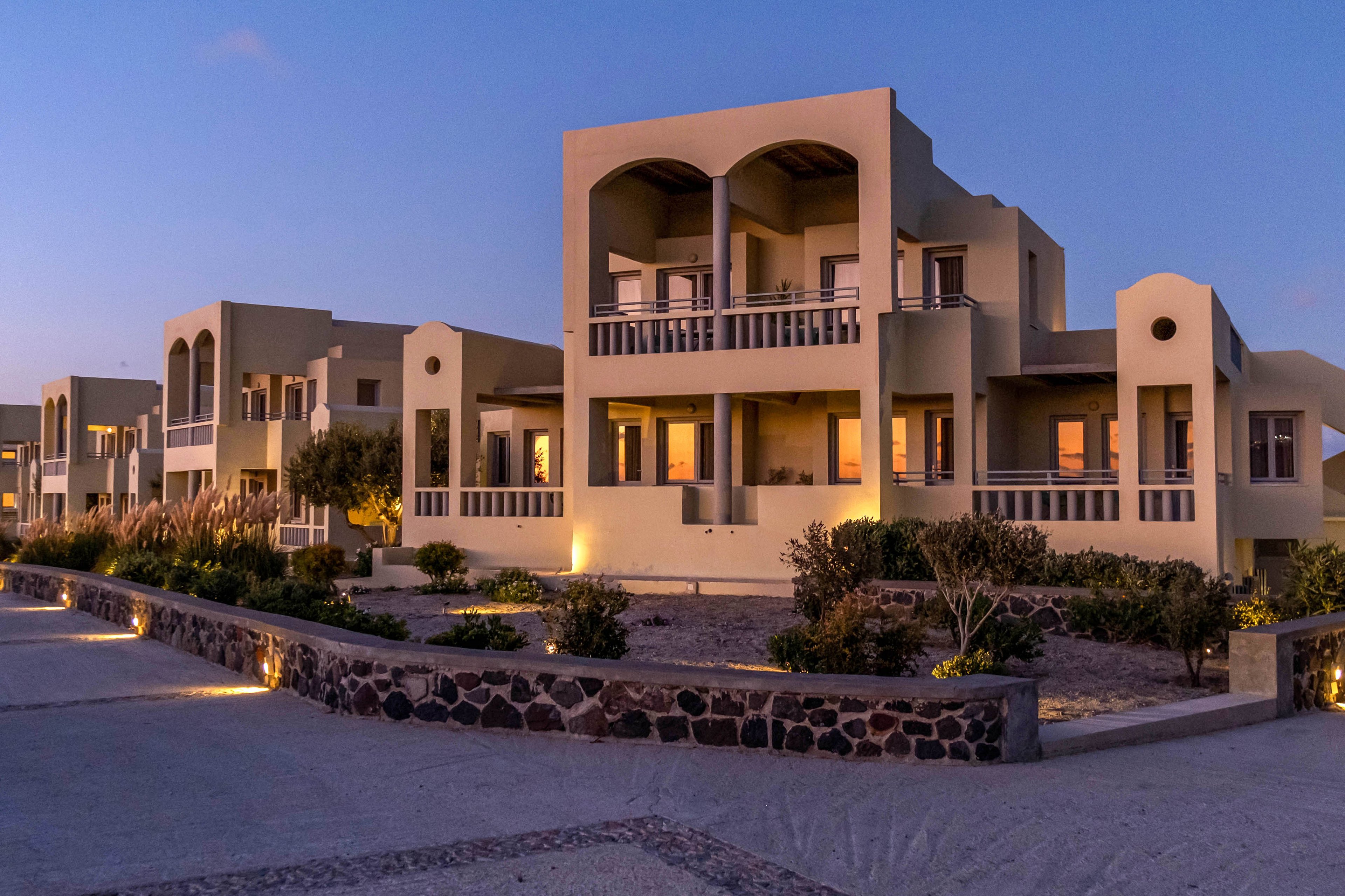 A row of modern beige villas is softly illuminated at dusk along a coastal hillside. Warm lights glow from the windows and pathway fixtures, contrasting with the cool blue evening sky. The architecture features arches, balconies, and clean geometric lines. The scene feels calm and serene, with no people visible and the sea faintly visible in the distance.