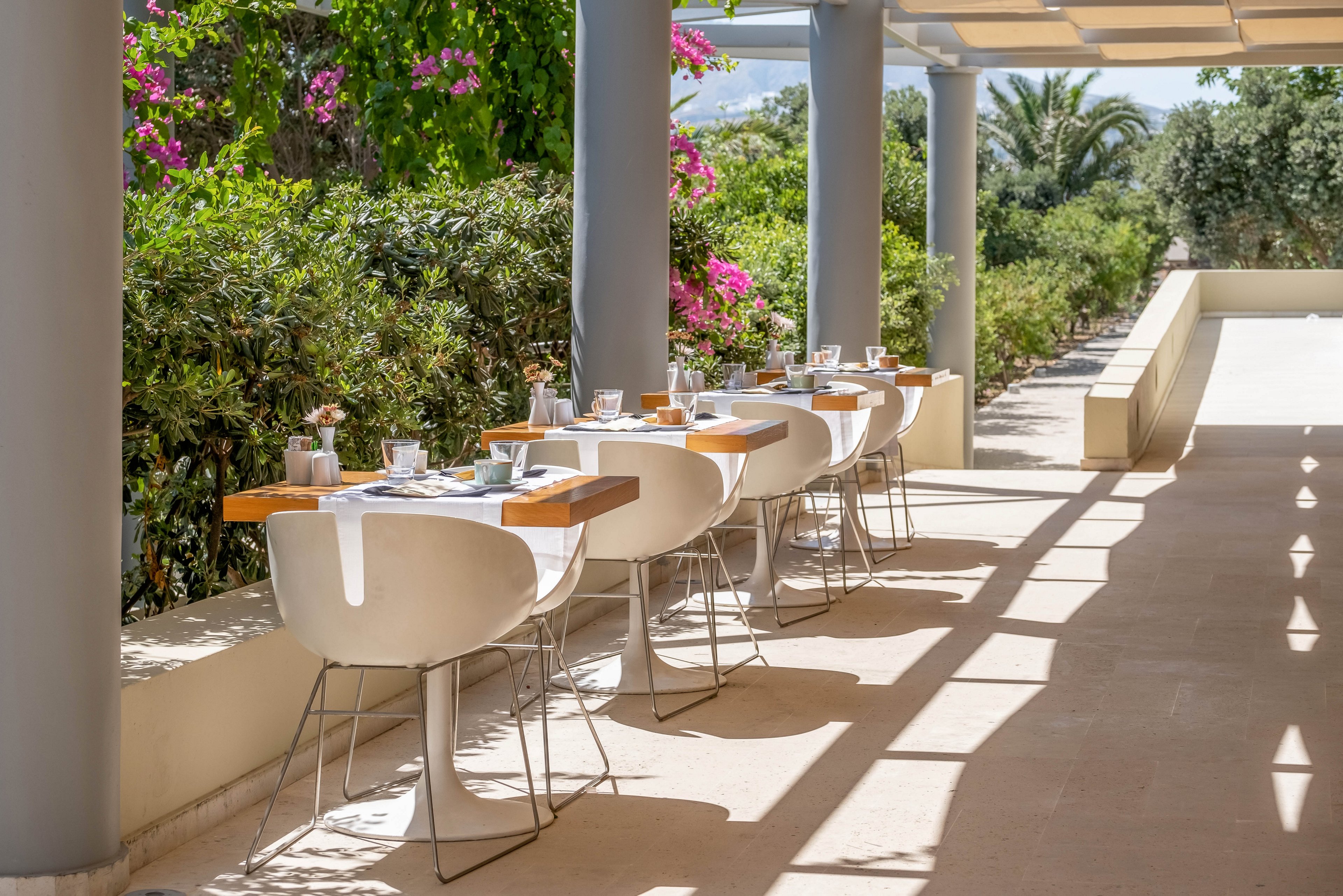 A row of modern white chairs and wooden tables is arranged neatly along a covered outdoor terrace. The tables are set with glassware, plates, and small floral arrangements, suggesting a prepared dining service. Sunlight filters through the pergola roof, casting shadows on the floor and creating a bright, inviting atmosphere. Lush greenery and flowering plants border the terrace, with palm trees visible in the background.