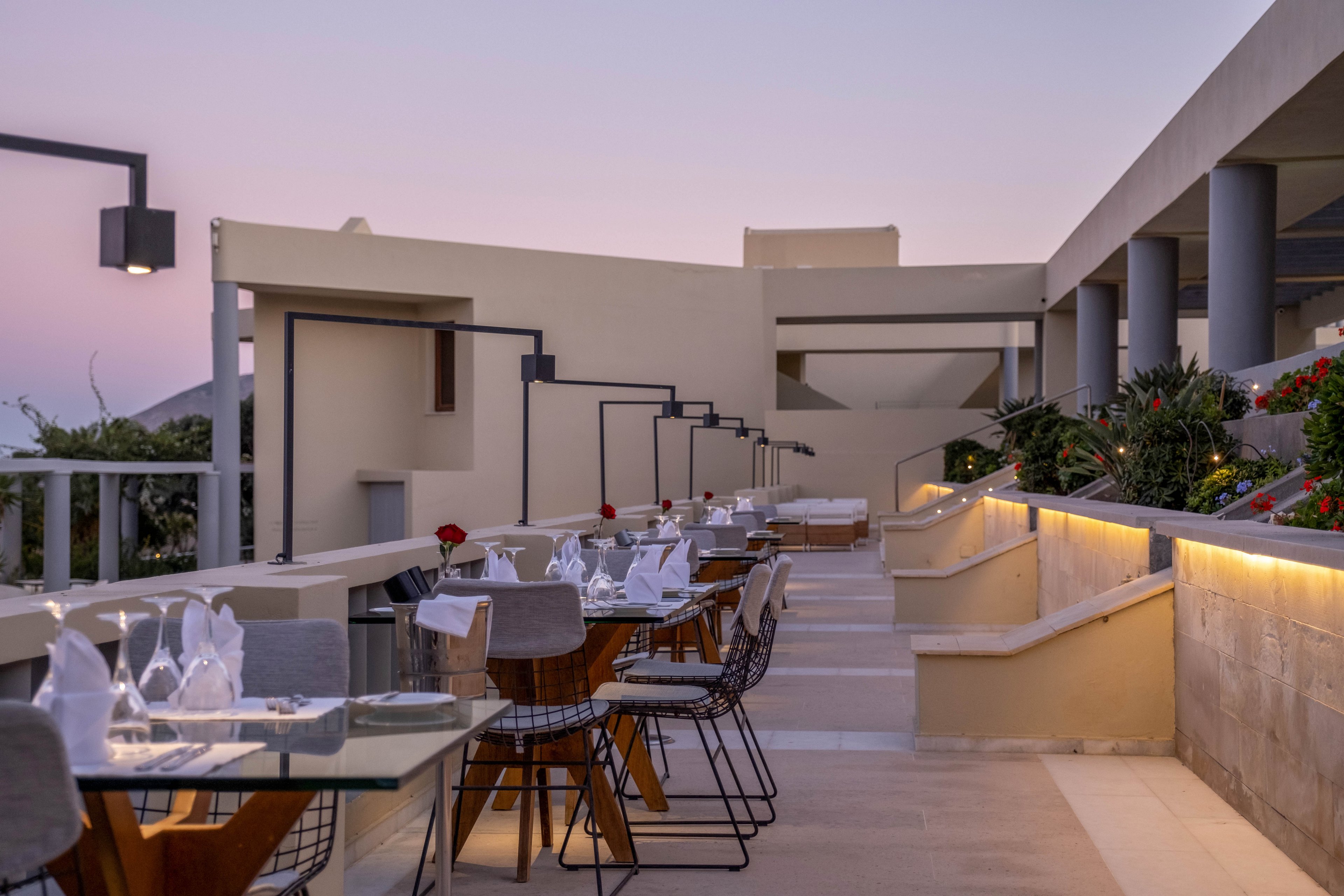 An elegant outdoor restaurant terrace is set for dinner on a modern rooftop. Glass-topped tables with neatly arranged place settings and folded napkins line a smooth stone walkway. Contemporary chairs and minimalist architectural lines create a stylish, upscale atmosphere. Soft lighting and a pastel evening sky enhance the tranquil, romantic mood of the scene.