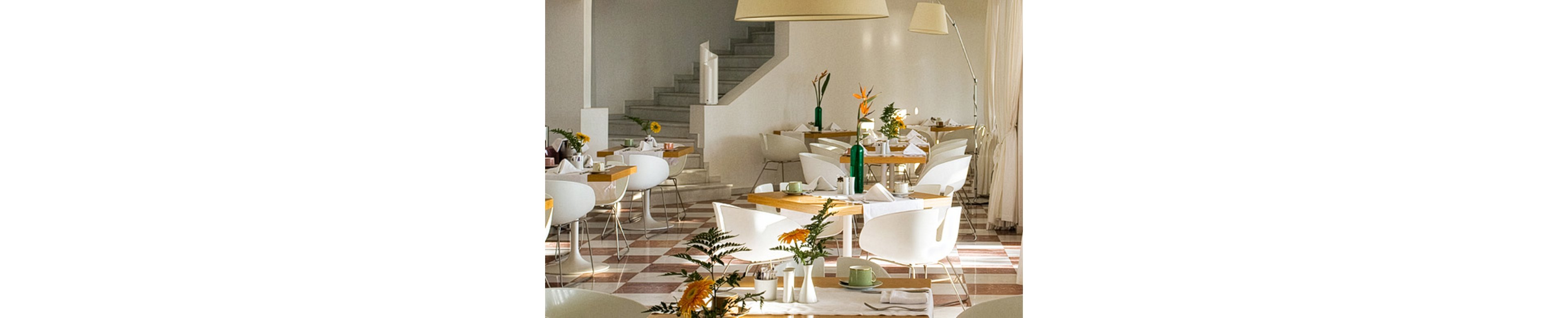 A bright, modern restaurant interior features white chairs and light wooden tables neatly arranged. Large windows with sheer curtains allow natural light to fill the space, creating an airy atmosphere. Decorative flowers, bottles, and tableware are placed on the tables, adding subtle color accents. A staircase in the background and contemporary floor lamps contribute to the clean, minimalist design.