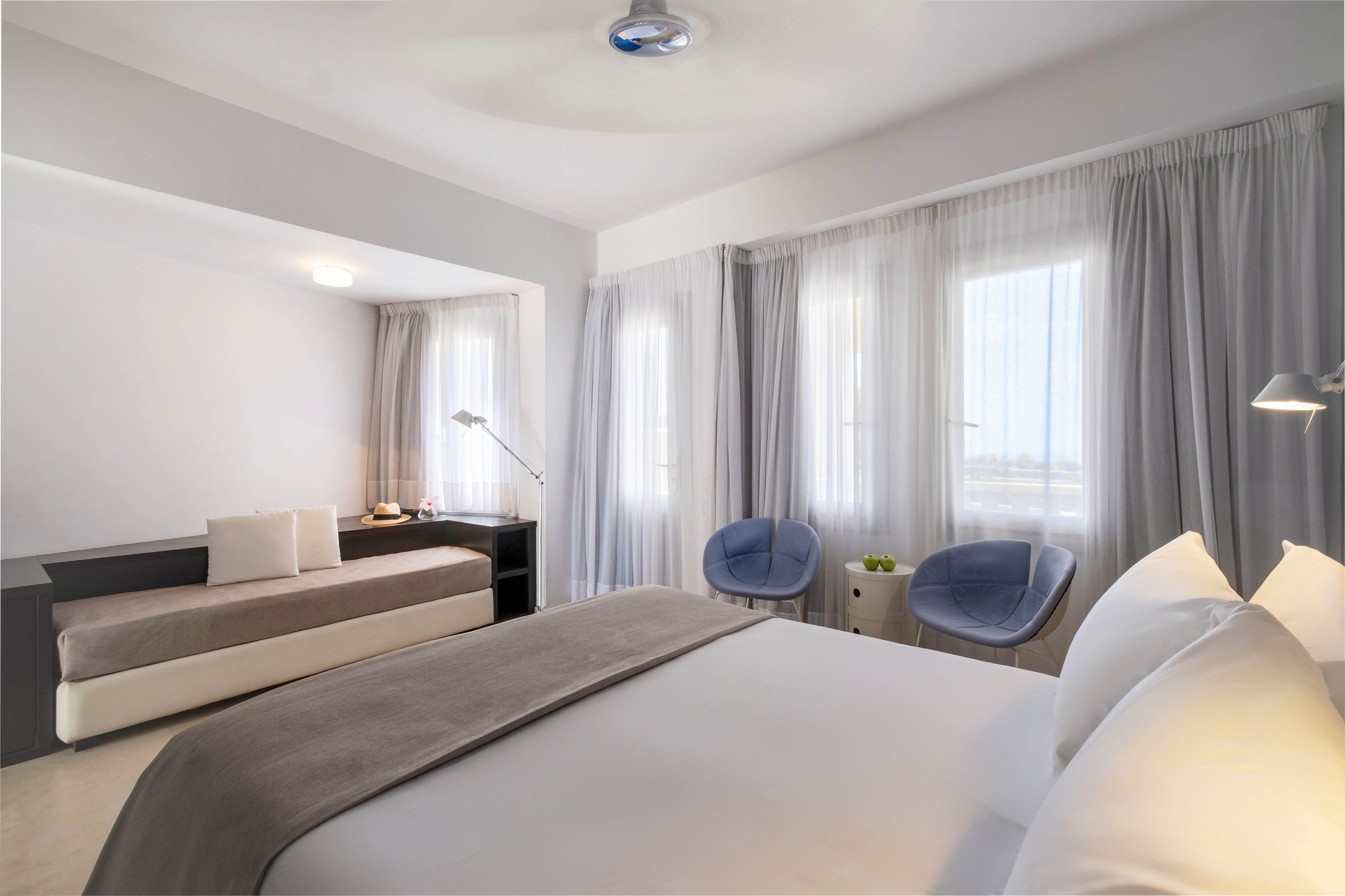 A contemporary bedroom features a large neatly made bed with white linens and a gray throw. The room includes a built-in daybed along the wall and a sleek black desk with a task lamp. Two modern blue chairs sit near tall windows covered with sheer curtains, allowing soft daylight to fill the space. Neutral tones and minimal decor create a calm, airy atmosphere suitable for a hotel or guest room.