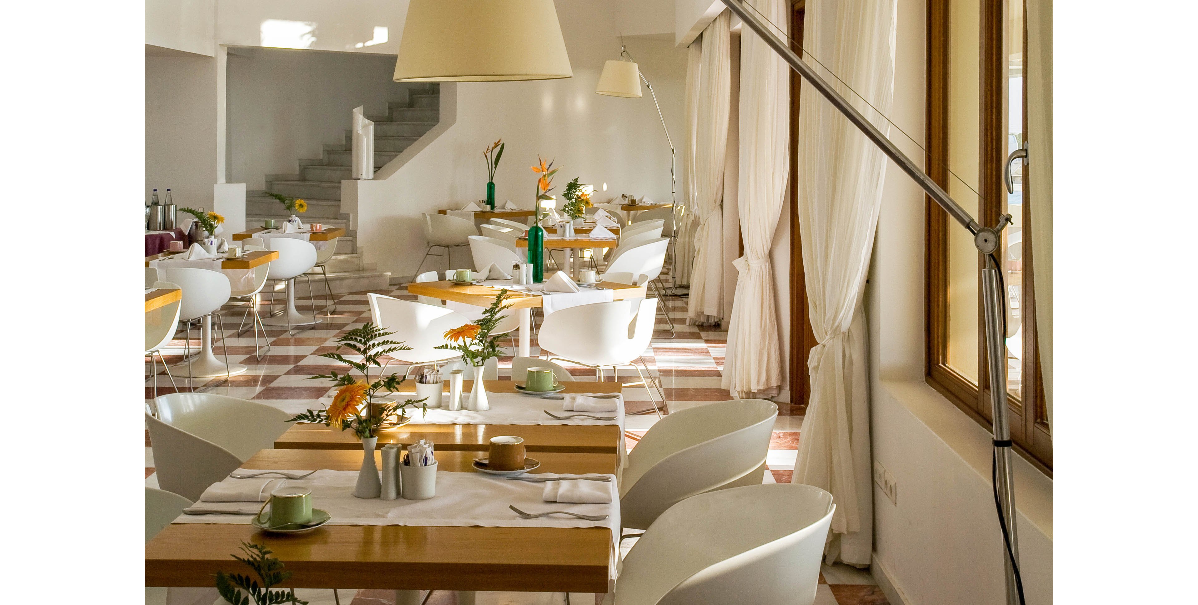 A contemporary dining room is filled with soft natural light streaming through large curtained windows. White chairs and light wooden tables are neatly arranged, each set with cups, plates, and small vases of yellow flowers. A large floor lamp with an extended arm adds a stylish design element to the space. The overall atmosphere feels airy, welcoming, and ideal for relaxed meals or casual gatherings.