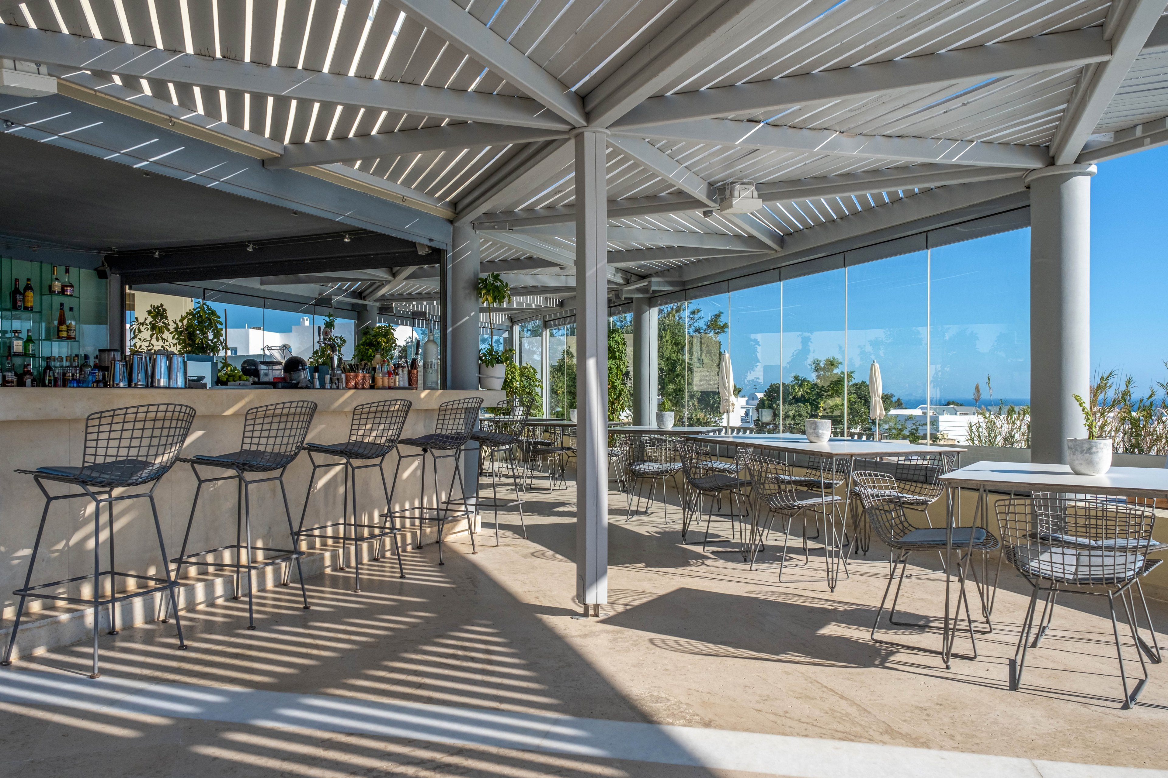 A contemporary open-air bar features high metal stools lined along a sleek counter. Adjacent tables and wire chairs sit under a geometric pergola that casts striped shadows across the floor. Large glass panels reveal a bright blue sky and distant sea, creating a relaxed coastal atmosphere. Potted plants and minimalist decor enhance the clean, modern aesthetic of the space.
