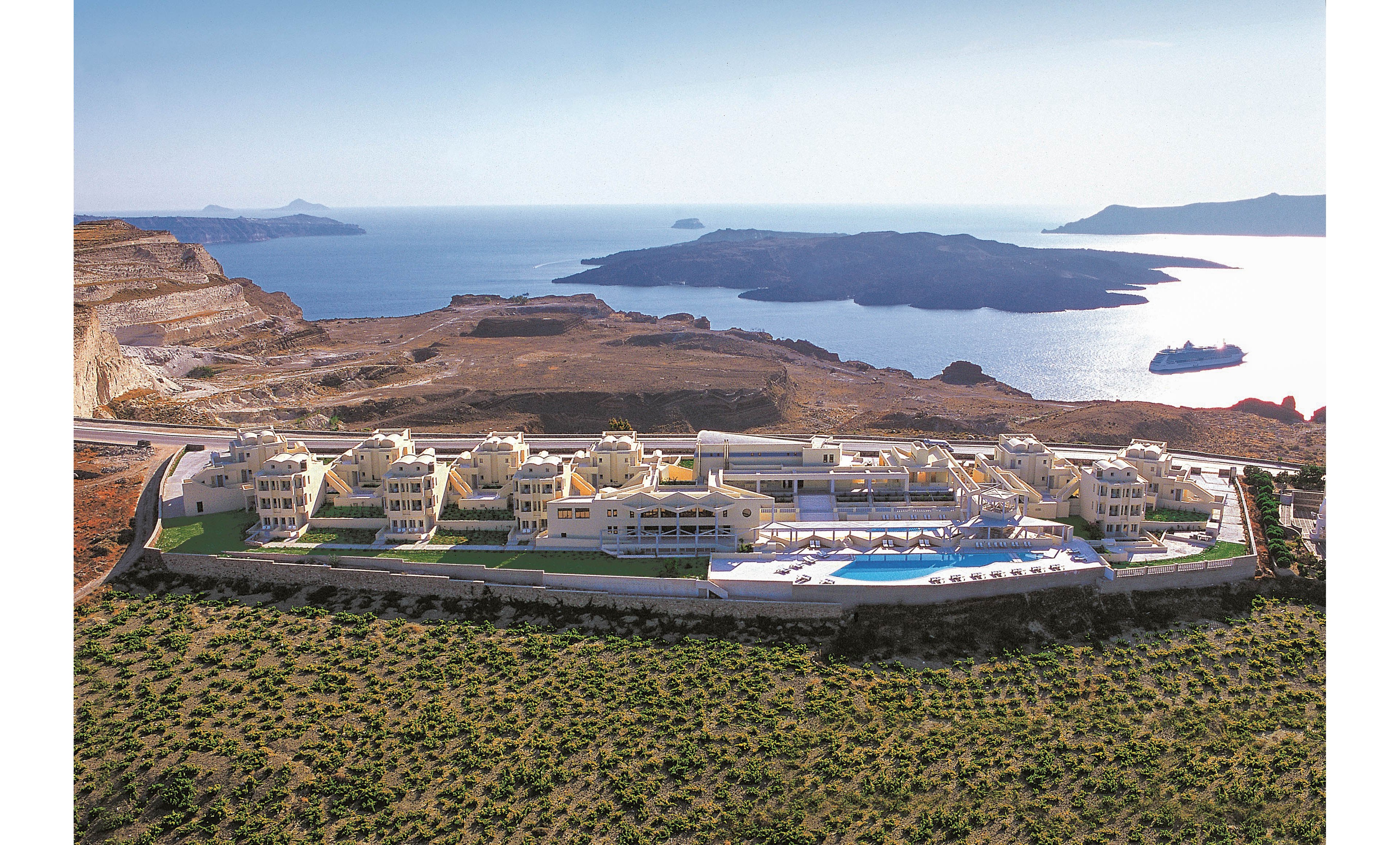 A large resort complex with multiple cream-colored buildings sits dramatically on a clifftop above the sea. The property features a prominent outdoor swimming pool and sun terrace facing the water. Rugged rocky terrain and sparse vegetation surround the resort, contrasting with the deep blue ocean and distant islands. The scene suggests a tranquil, upscale vacation destination in a coastal or island setting.