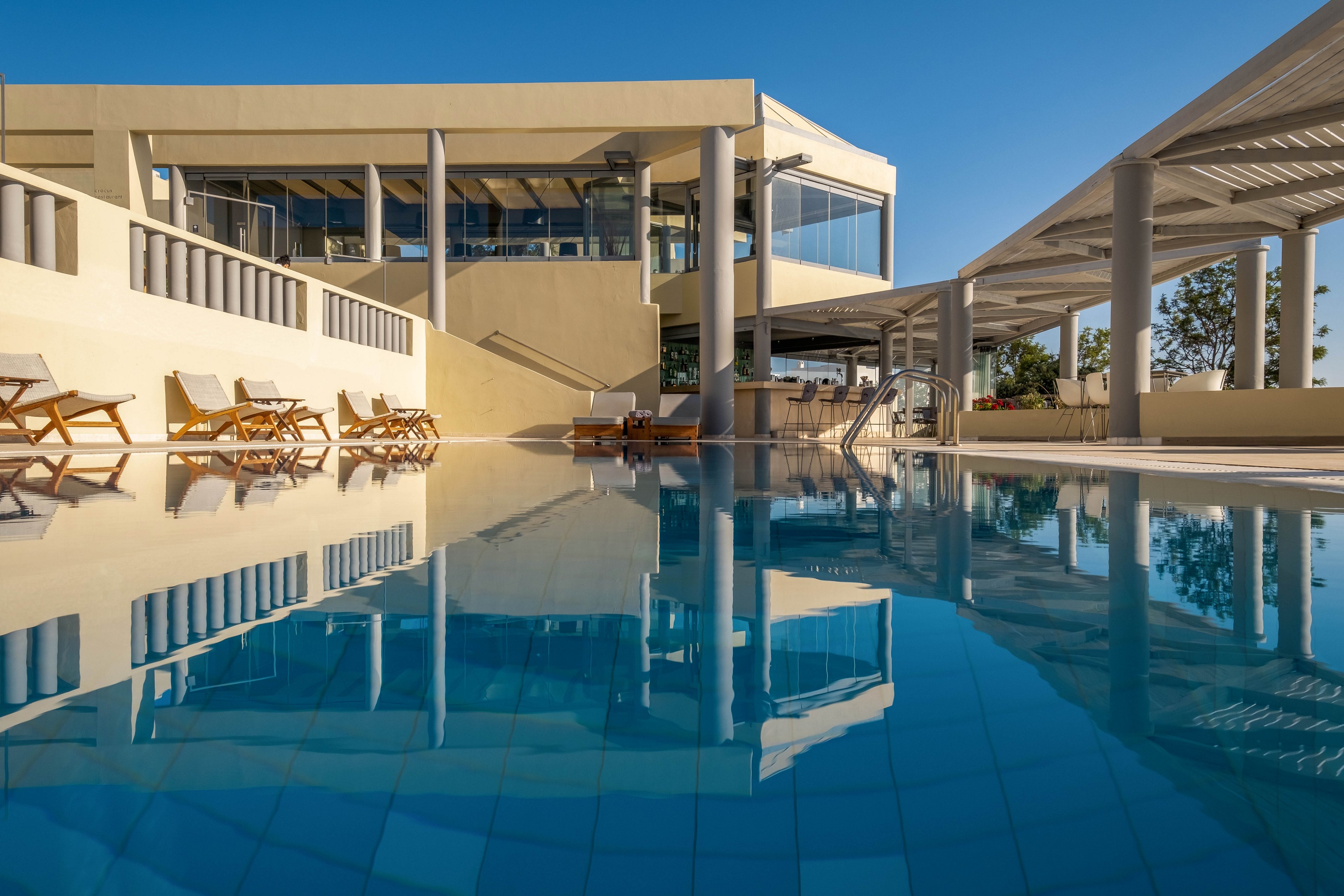 A modern resort building overlooks a clear blue swimming pool under a bright morning sky. Wooden lounge chairs line the poolside terrace, suggesting a relaxing leisure setting. The architecture features clean lines, large glass windows, and shaded pergola structures. Calm reflections of the building and sky appear on the still water, creating a serene and inviting atmosphere.