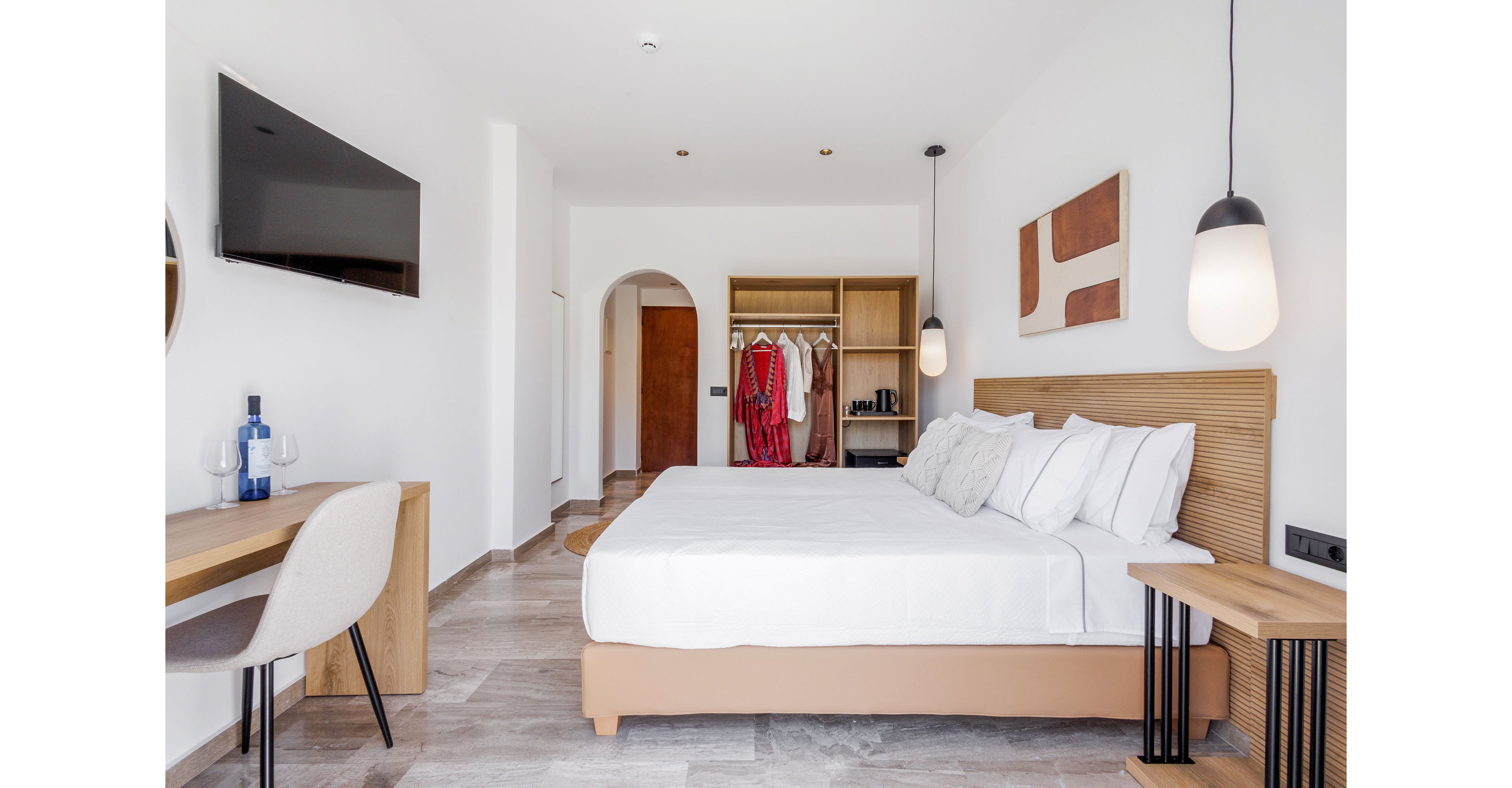 A contemporary hotel room features a large bed with white linens and multiple pillows against a wooden headboard. An open wardrobe displays hanging clothes and shelves, set near an arched doorway. A wall-mounted flat-screen TV faces the bed, while a wooden desk with a chair and a bottle with two glasses creates a small workspace. Minimalist pendant lighting and abstract wall art enhance the clean, modern aesthetic of the bright interior.