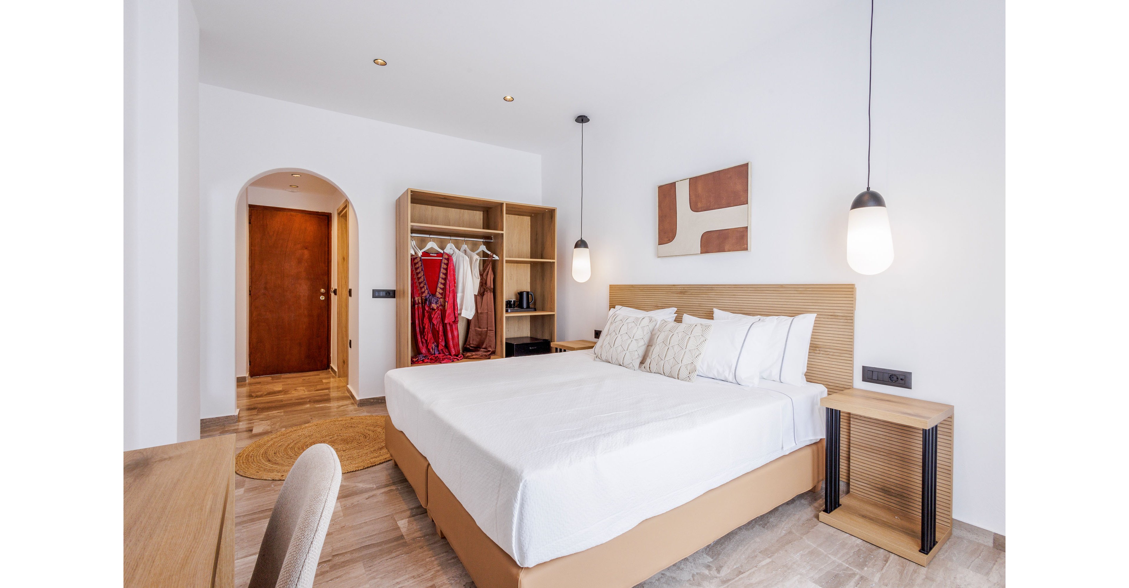 A contemporary bedroom features a neatly made double bed with white linens and decorative pillows. An open wooden wardrobe displays hanging clothes and accessories near an arched doorway. Minimalist pendant lights and abstract wall art add a modern touch to the clean, white interior. The space feels airy and inviting, with neutral tones and natural textures throughout.