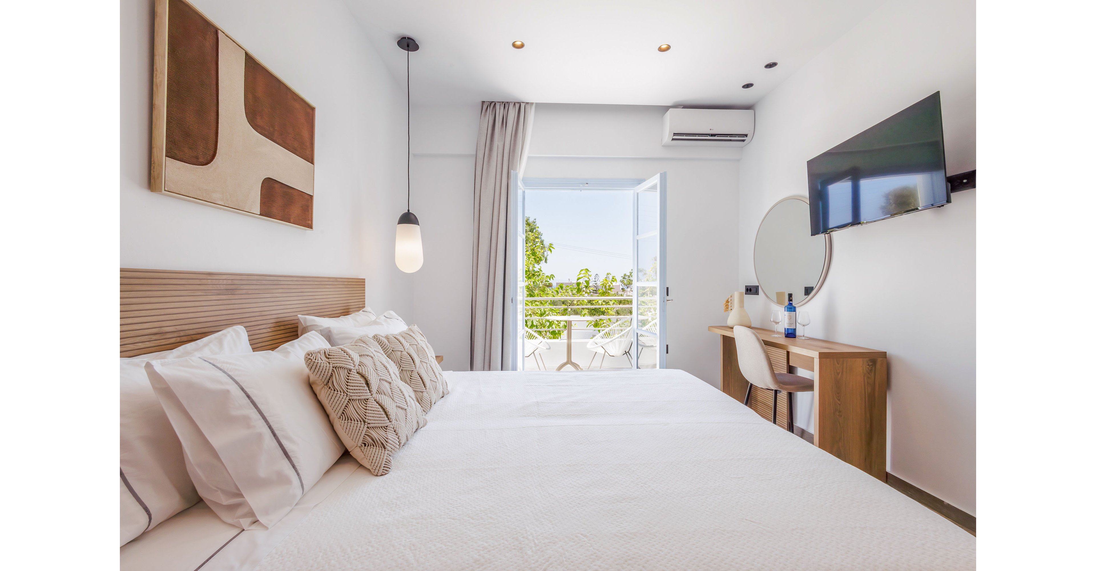 A modern, minimalist bedroom features a neatly made double bed with textured cushions and a wooden headboard. The room is decorated in white tones with simple wall art, a hanging pendant light, and a mounted flat-screen TV. A small wooden desk with a round mirror and chair sits beside the bed, holding a bottle and glass. Sheer curtains frame open balcony doors that reveal greenery and outdoor seating, filling the space with natural light.