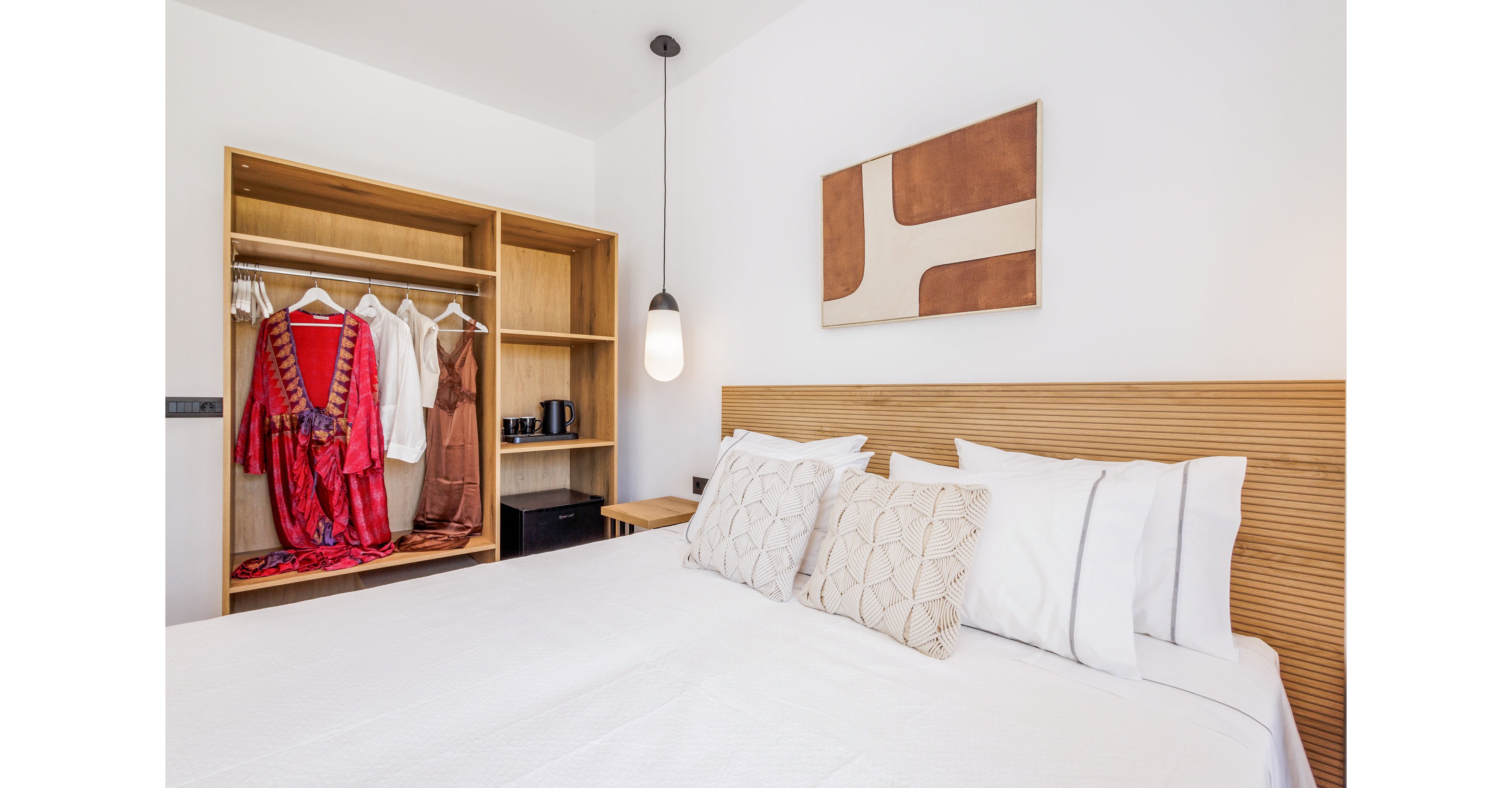 A neatly made double bed with white linens and patterned cushions sits in a modern, minimalist bedroom. An open wooden wardrobe displays hanging clothes, including a bright red dress and white garments, alongside shelves holding accessories and a kettle. A pendant light hangs beside the bed, and abstract wall art adds a contemporary touch to the clean white walls. The overall scene feels tidy, inviting, and stylishly designed for a comfortable stay.