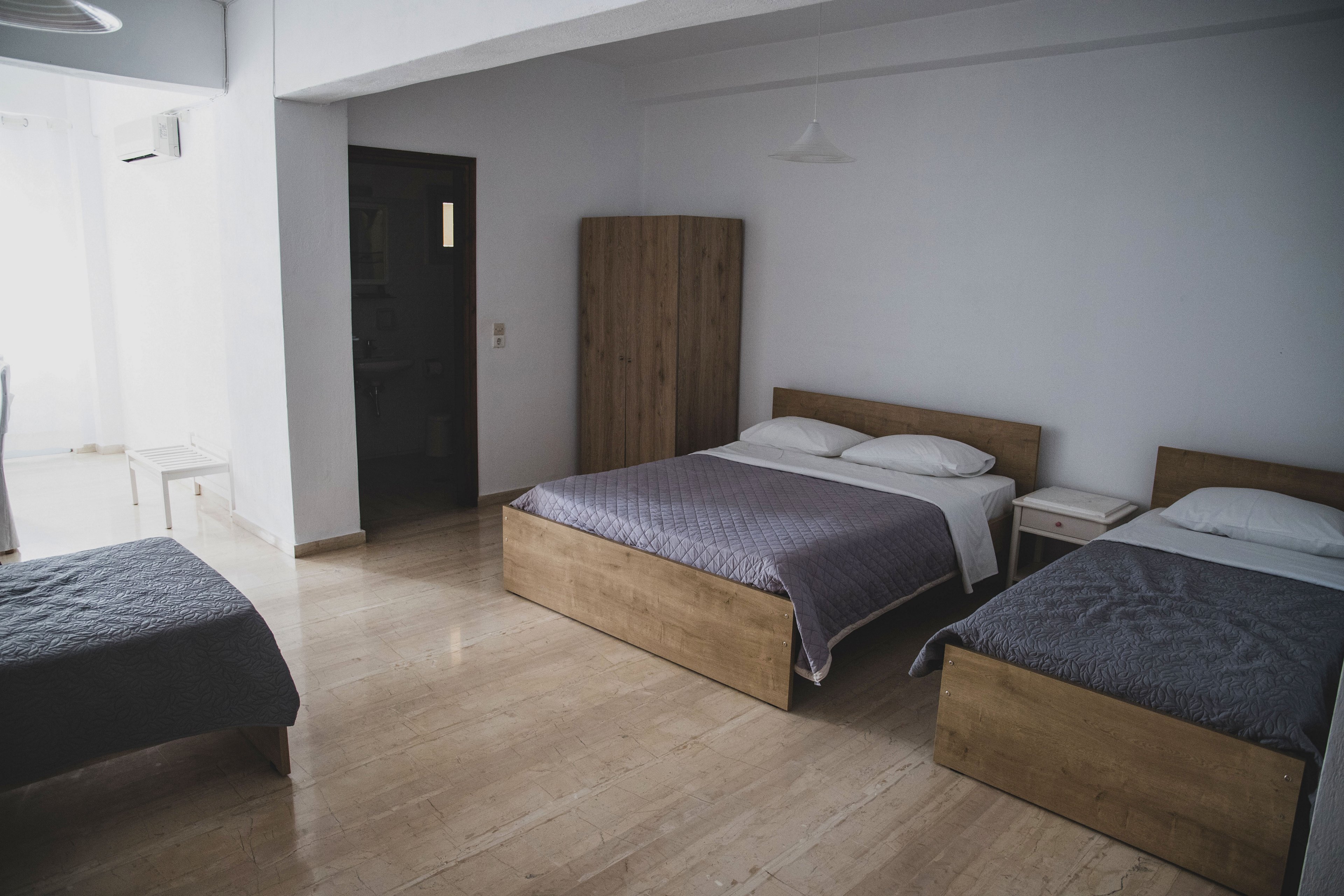 A simple, modern bedroom features three wooden beds with gray bedspreads and white pillows. The room has light-colored flooring and white walls, creating a clean and minimalist atmosphere. A wardrobe and bedside tables are placed against the wall near a dark door. Soft daylight enters from a large window, enhancing the calm and neutral color palette.