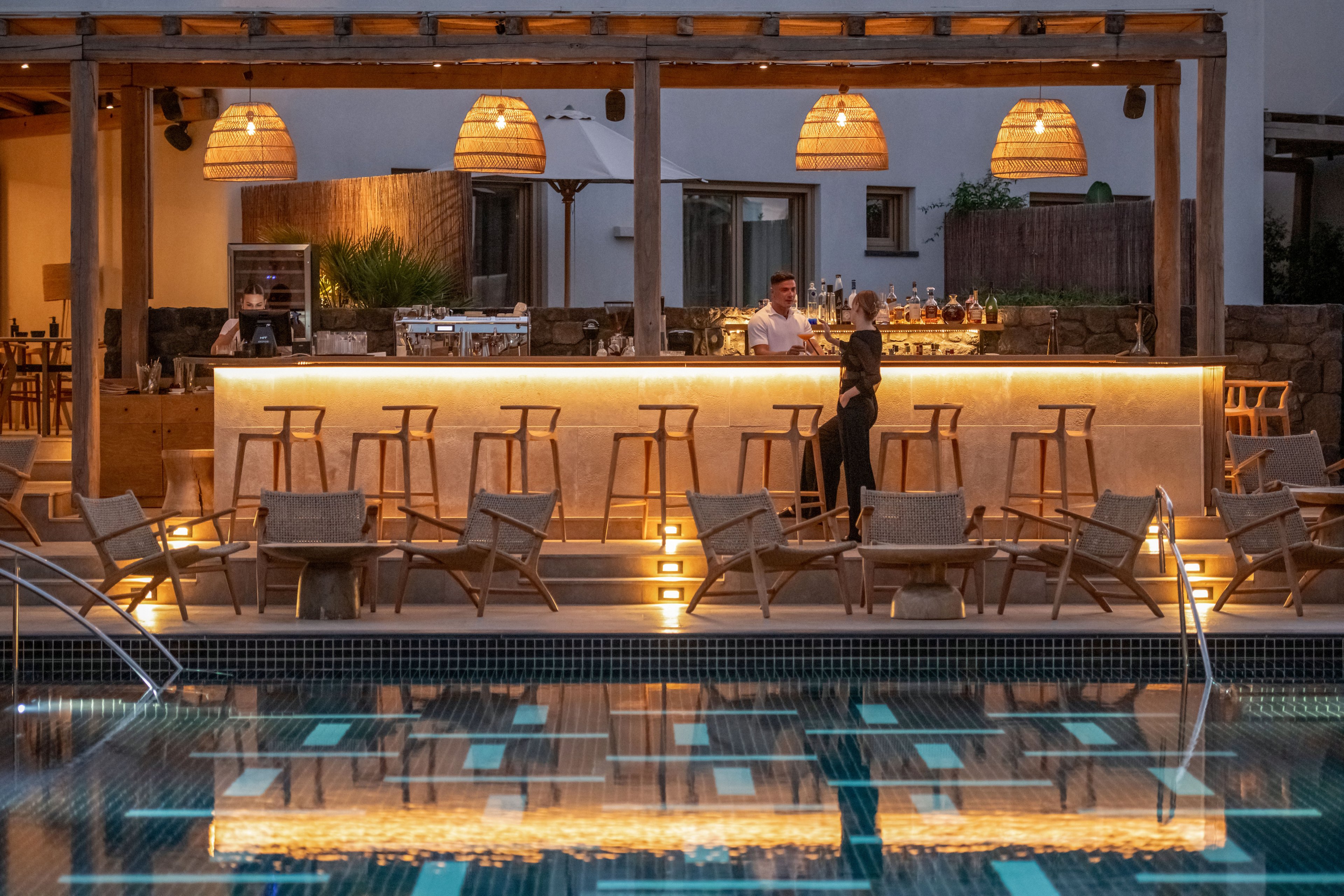An illuminated outdoor bar stands beside a calm swimming pool in the early evening. Wooden stools and lounge chairs line the bar, with two people conversing near the counter. Warm pendant lights and under-counter lighting create a cozy, inviting atmosphere. The surrounding architecture features natural materials and neutral tones, enhancing the relaxed resort setting.