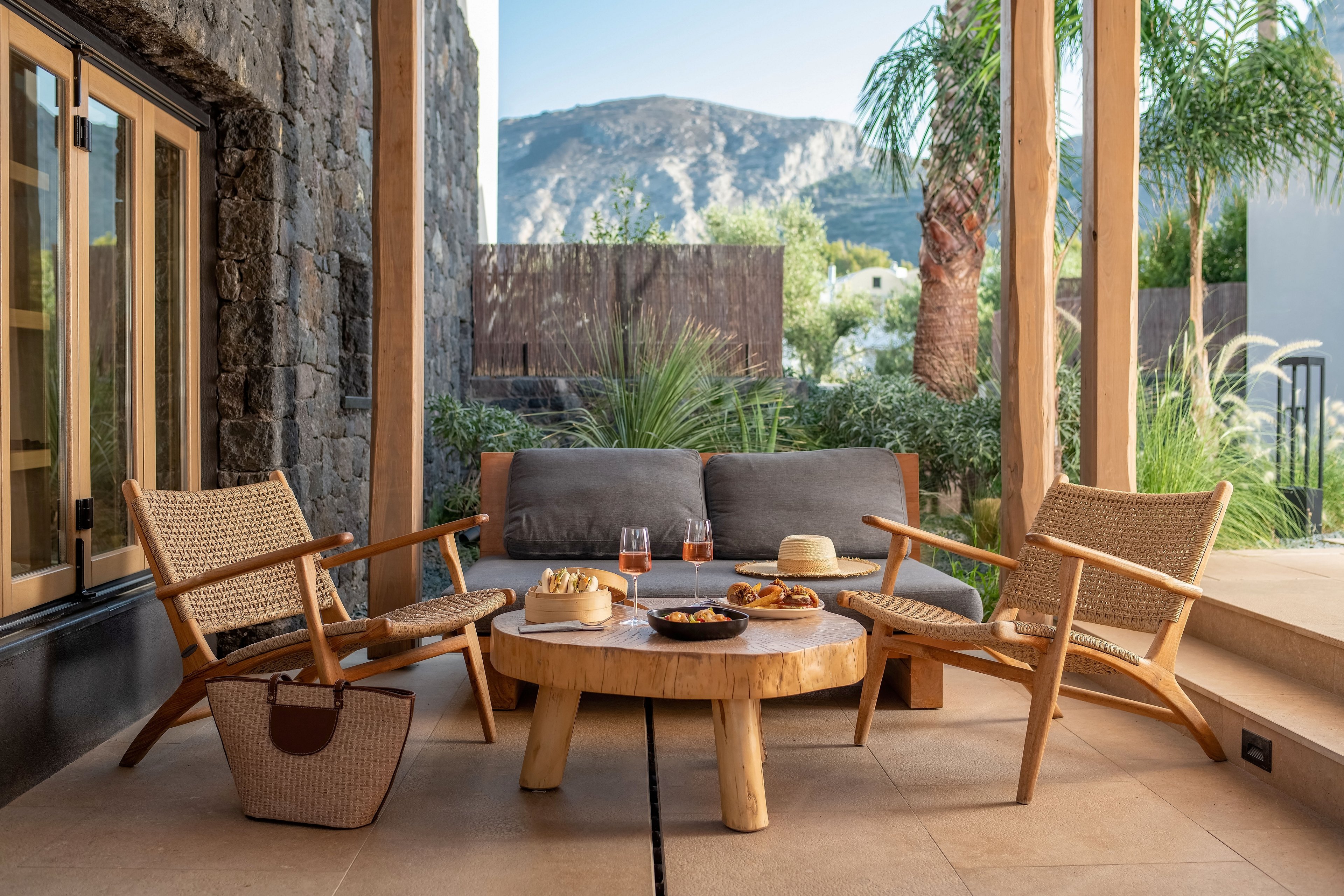 A modern outdoor patio features a wooden coffee table surrounded by woven lounge chairs and a cushioned sofa. On the table are plates of snacks and two drinks, suggesting a relaxed gathering. A straw hat and a handbag add a casual vacation feel to the scene. The space is framed by stone and wood architecture, lush greenery, and distant mountains under a clear sky.