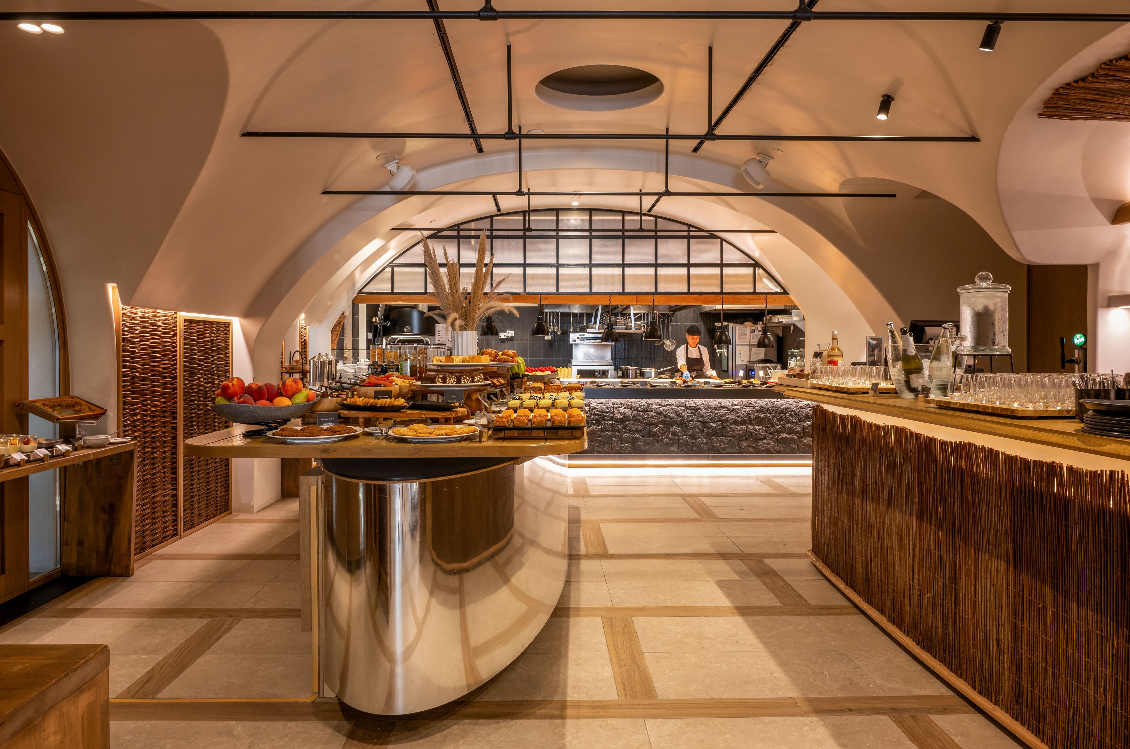 A contemporary buffet area is set inside an arched, stylish interior with warm lighting. Various pastries, breads, and fresh fruits are arranged on sleek wooden and metallic counters. In the background, an open kitchen with staff preparing food is visible behind a stone-front counter. The overall atmosphere feels inviting and upscale, combining natural materials with modern design elements.