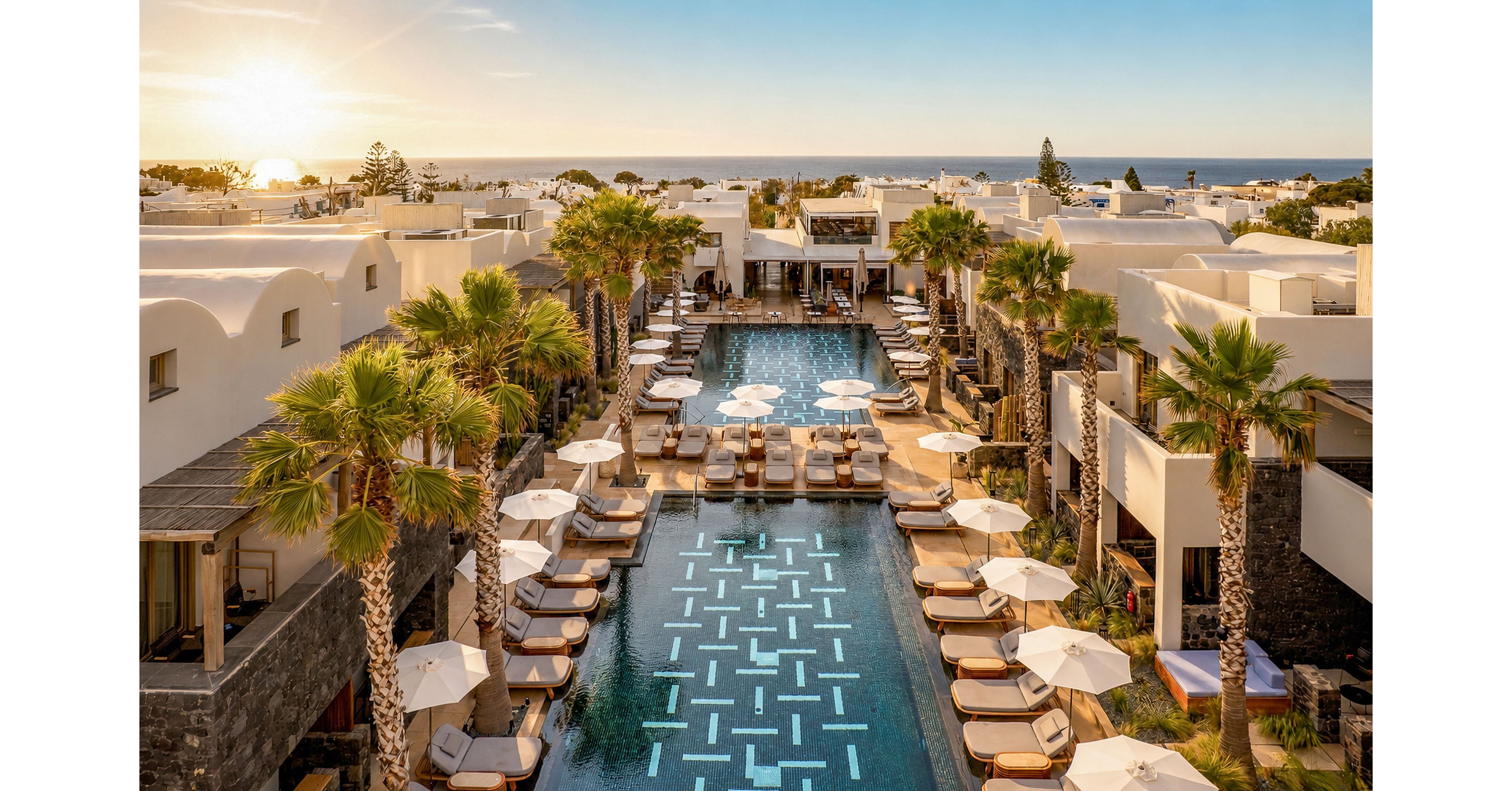 A luxurious resort features a long central swimming pool lined with sun loungers and umbrellas. White, low-rise buildings with curved roofs surround the pool, creating a modern Mediterranean-style courtyard. Tall palm trees add tropical greenery along the water’s edge, while warm golden sunlight illuminates the scene. In the distance, the sun sets over the sea, enhancing the tranquil and upscale vacation atmosphere.