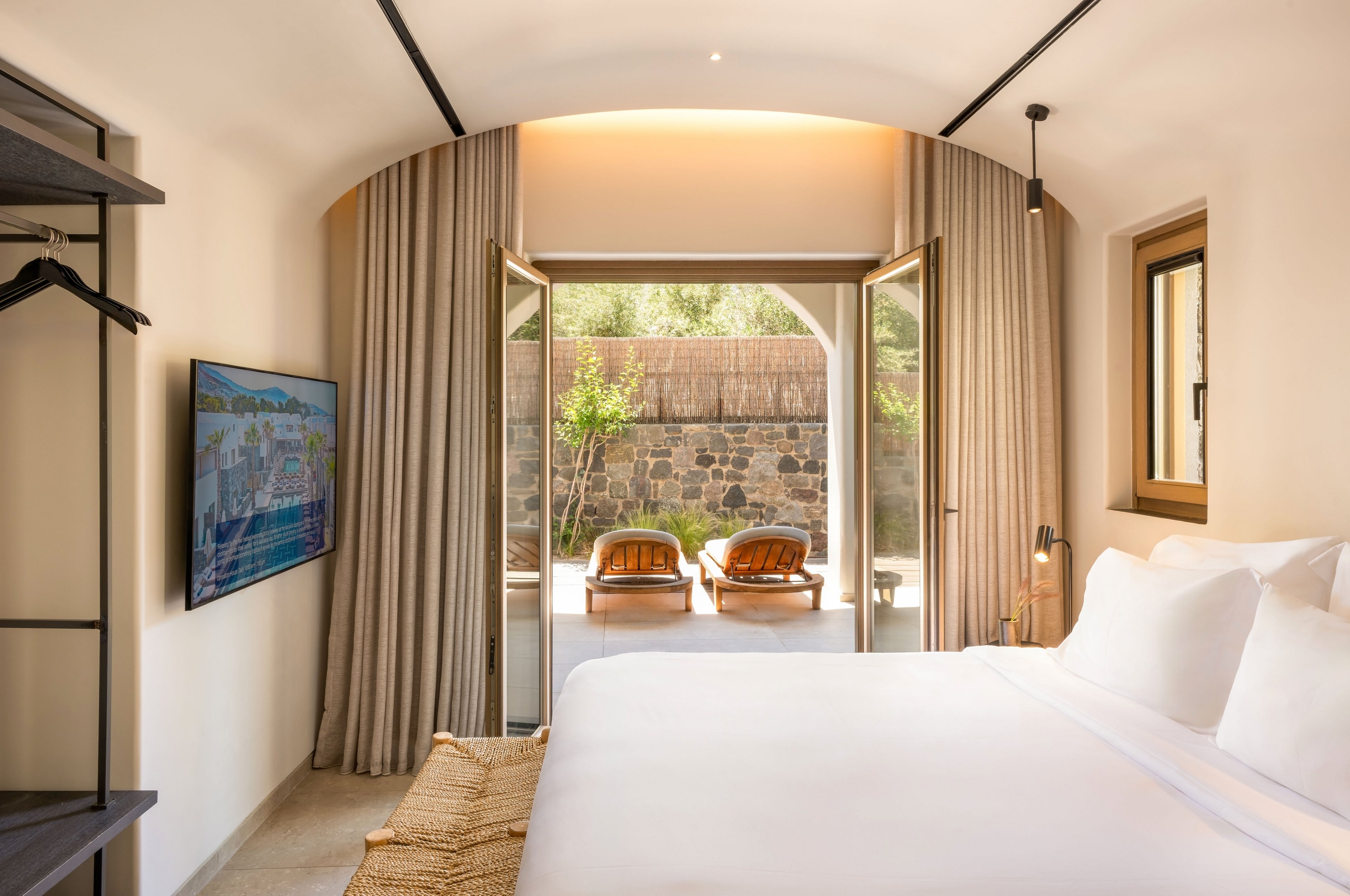 Contemporary hotel bedroom with a large white bed faces open glass doors leading to a sunny patio. Neutral-toned curtains frame the doorway, revealing two lounge chairs and greenery outside against a stone wall. A wall-mounted television and minimalist shelving add modern comfort to the space. Warm natural light and soft colors create a calm, inviting atmosphere ideal for relaxation.