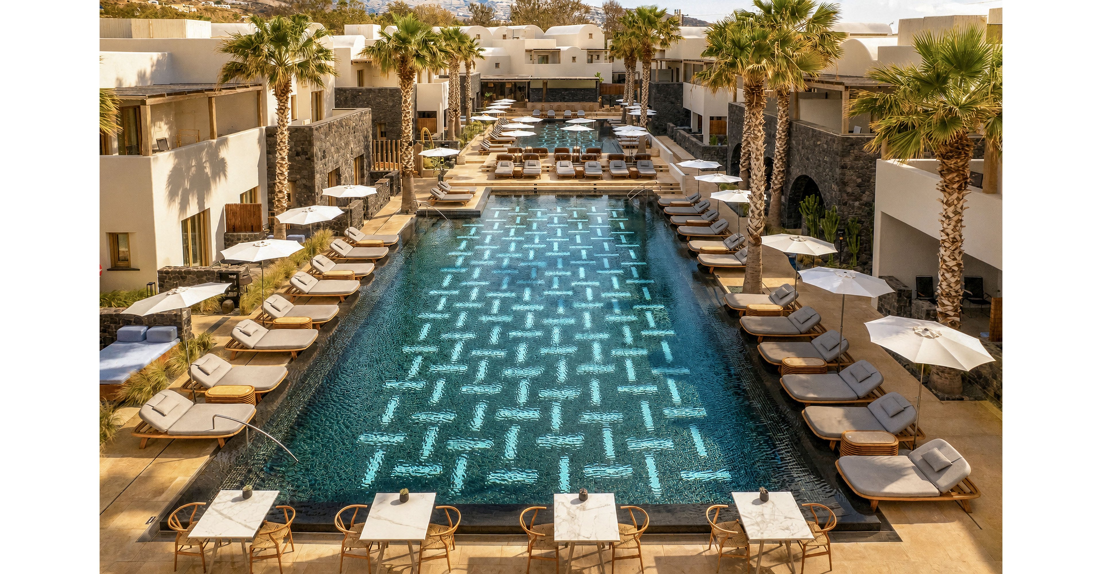 A large rectangular swimming pool is centered in a modern resort courtyard, surrounded by rows of cushioned sun loungers and umbrellas. Palm trees line the poolside, adding a tropical feel to the contemporary architecture of the surrounding buildings. Tables and chairs are arranged at the near end of the pool, suggesting an outdoor dining or lounge area. The scene is sunlit and tranquil, evoking a relaxed vacation atmosphere.