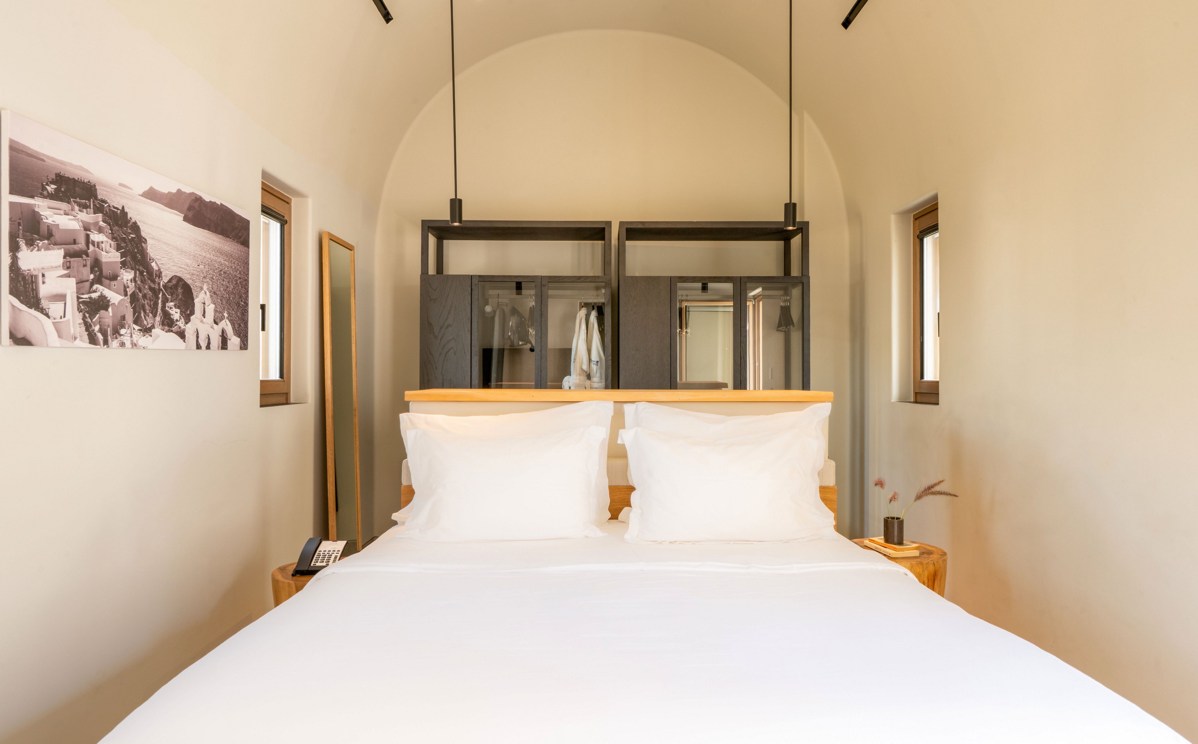 A neatly made bed with crisp white linens is centered in a minimalist hotel room. The space features light-colored walls, two small side tables, and narrow windows on each side of the bed. At the back of the room, a modern open wardrobe and bathroom area are visible through glass partitions. A black-and-white landscape photograph hangs on the left wall, adding a subtle decorative touch to the clean, contemporary interior.