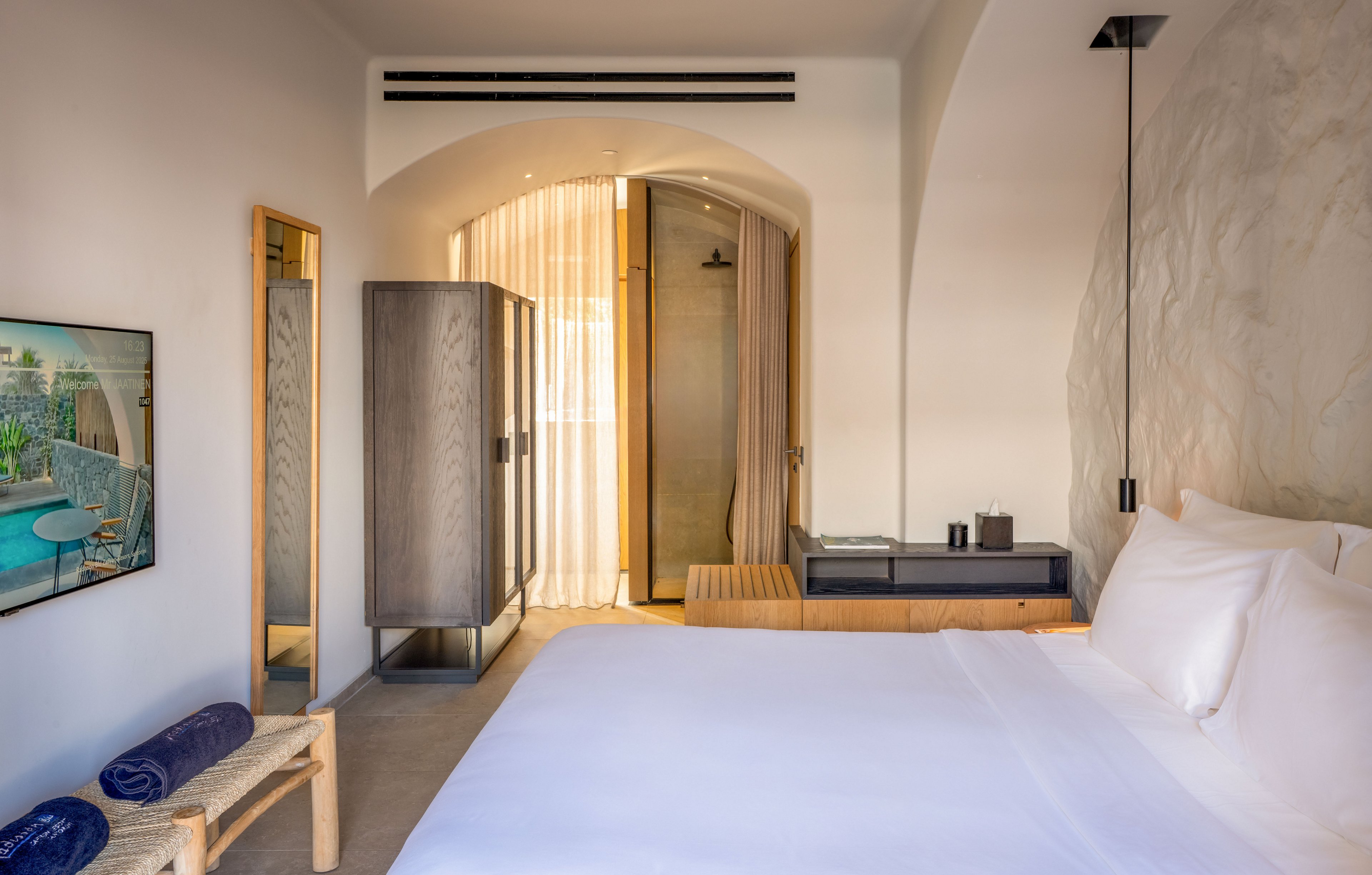 A bright, modern hotel bedroom features a neatly made white bed in the foreground. The room includes a wooden bench with rolled towels, a tall mirror, and a sleek wardrobe beside a console table. Sheer curtains cover an arched doorway leading to a bathroom, adding softness to the clean architectural lines. A wall-mounted television displays an outdoor balcony scene, enhancing the airy, contemporary atmosphere.