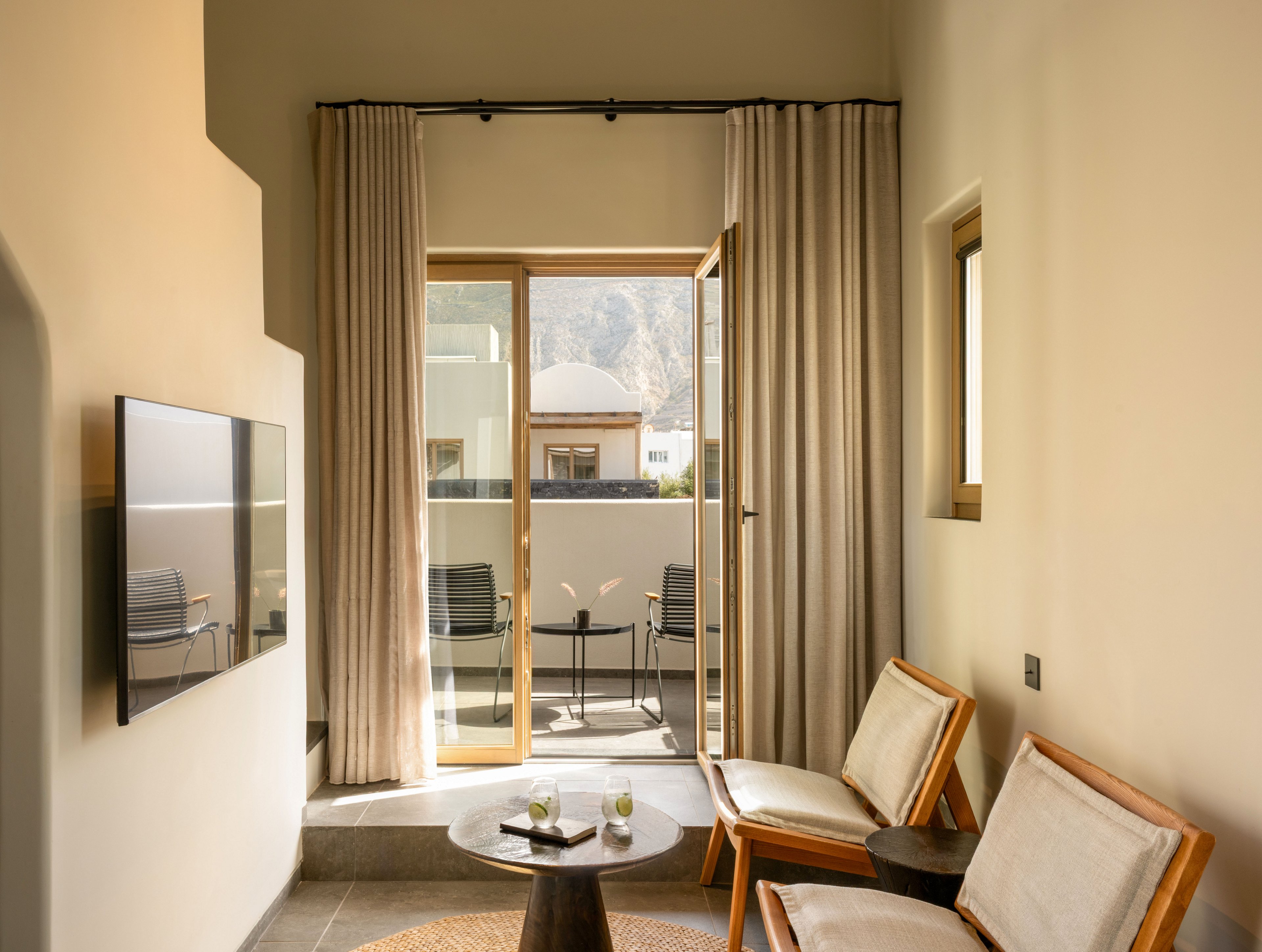A modern hotel room features two cushioned wooden chairs and a small round table with drinks and snacks. Floor-to-ceiling curtains frame a glass door leading to a balcony with outdoor chairs and a mountain view. A flat-screen television is mounted on the wall opposite the seating area, and soft neutral tones create a calm atmosphere. Natural light fills the space, highlighting the minimalist decor and comfortable setting.
