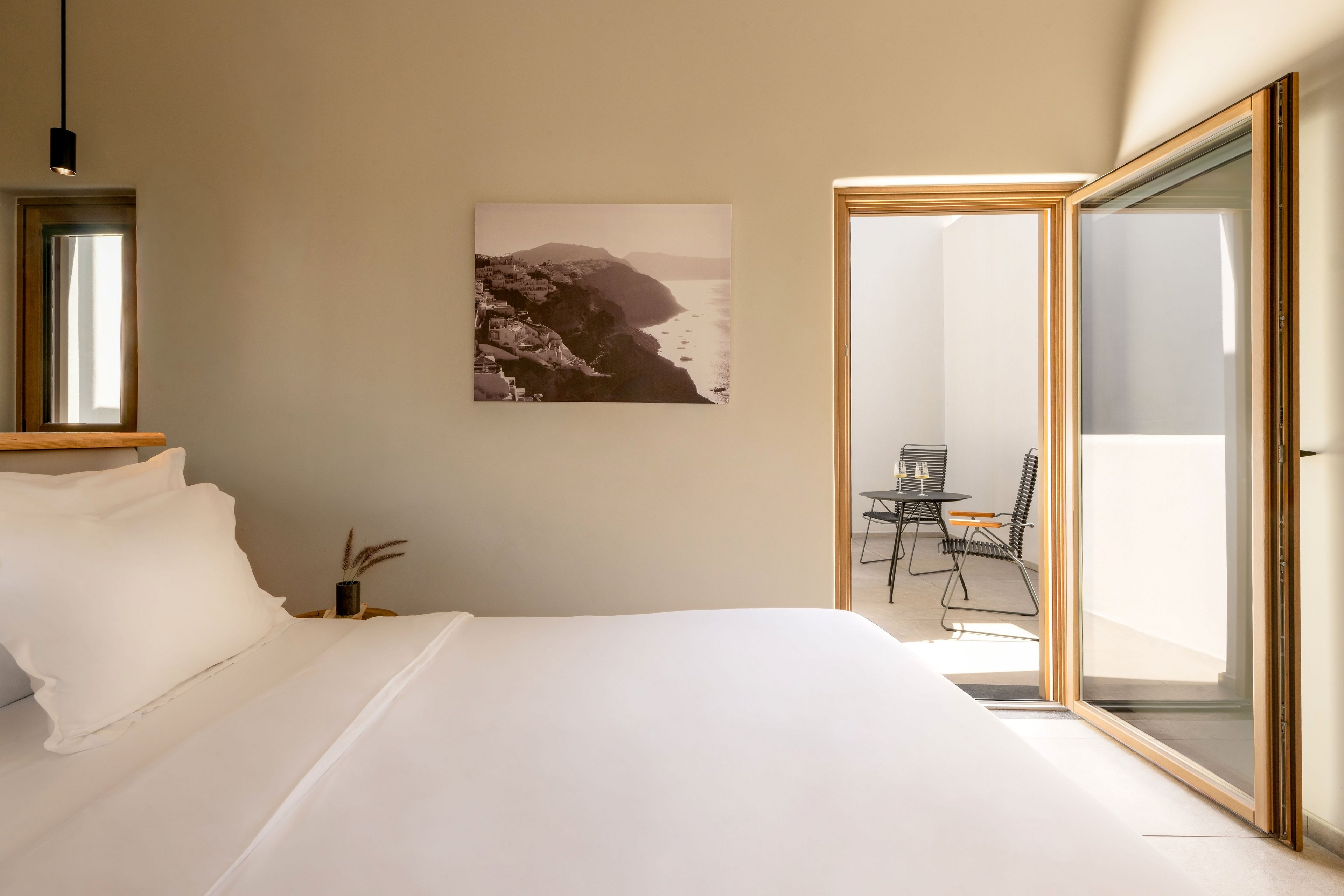 A neatly made bed with crisp white linens dominates a minimalist bedroom. Warm beige walls and a framed coastal photograph create a calm, contemporary atmosphere. An open sliding glass door leads to a small terrace with two chairs and a table in bright sunlight. The overall scene suggests a serene vacation or boutique hotel stay.