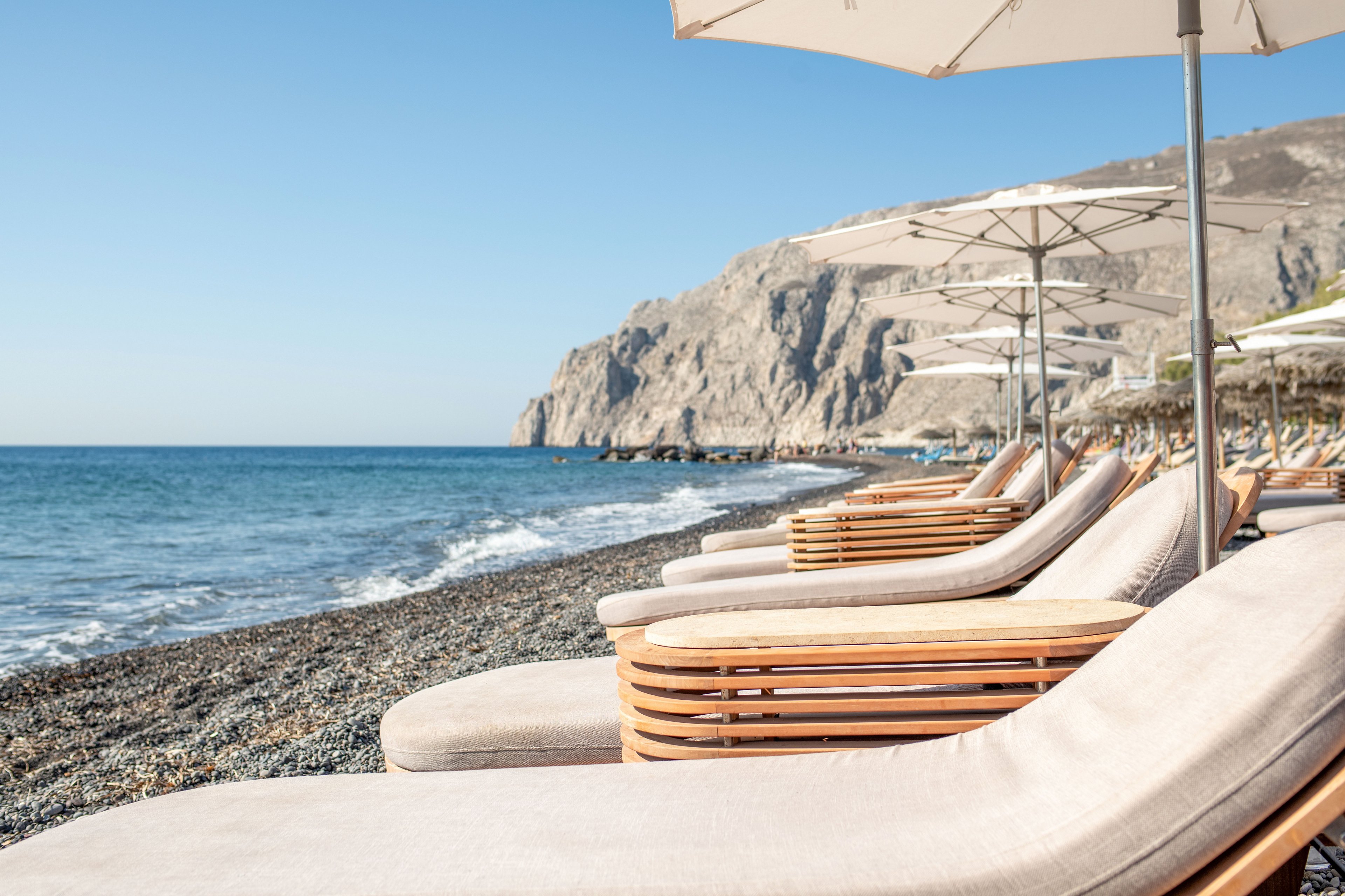 A row of cushioned wooden sun loungers sits under white umbrellas along a rocky beach. Calm blue waves gently meet the shoreline, with a rugged mountain range rising in the background. The clear sky and soft neutral tones create a serene, minimalist coastal atmosphere. The scene suggests a peaceful vacation setting focused on relaxation by the sea.