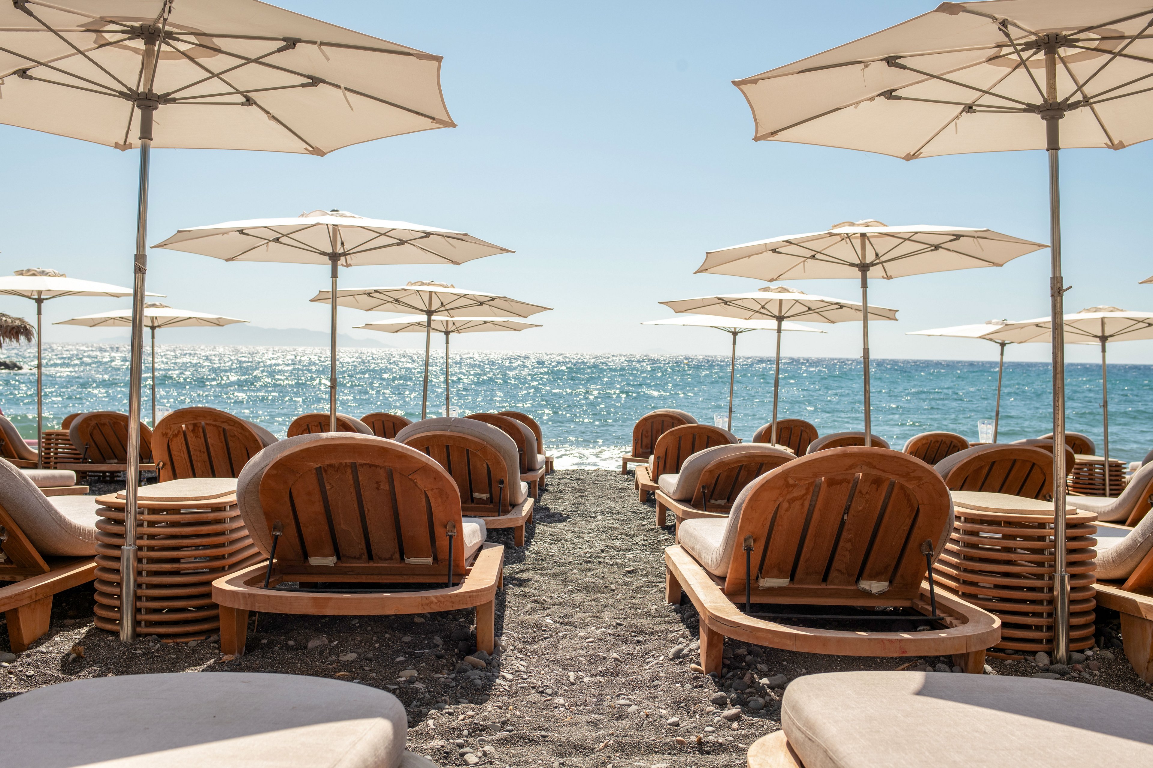 A row of wooden sun loungers is arranged on a sandy beach facing the sea. Large white umbrellas provide shade over the chairs, creating a resort-like setting. The calm blue water sparkles in the sunlight under a clear sky. The scene suggests a relaxing summer holiday atmosphere by the ocean.