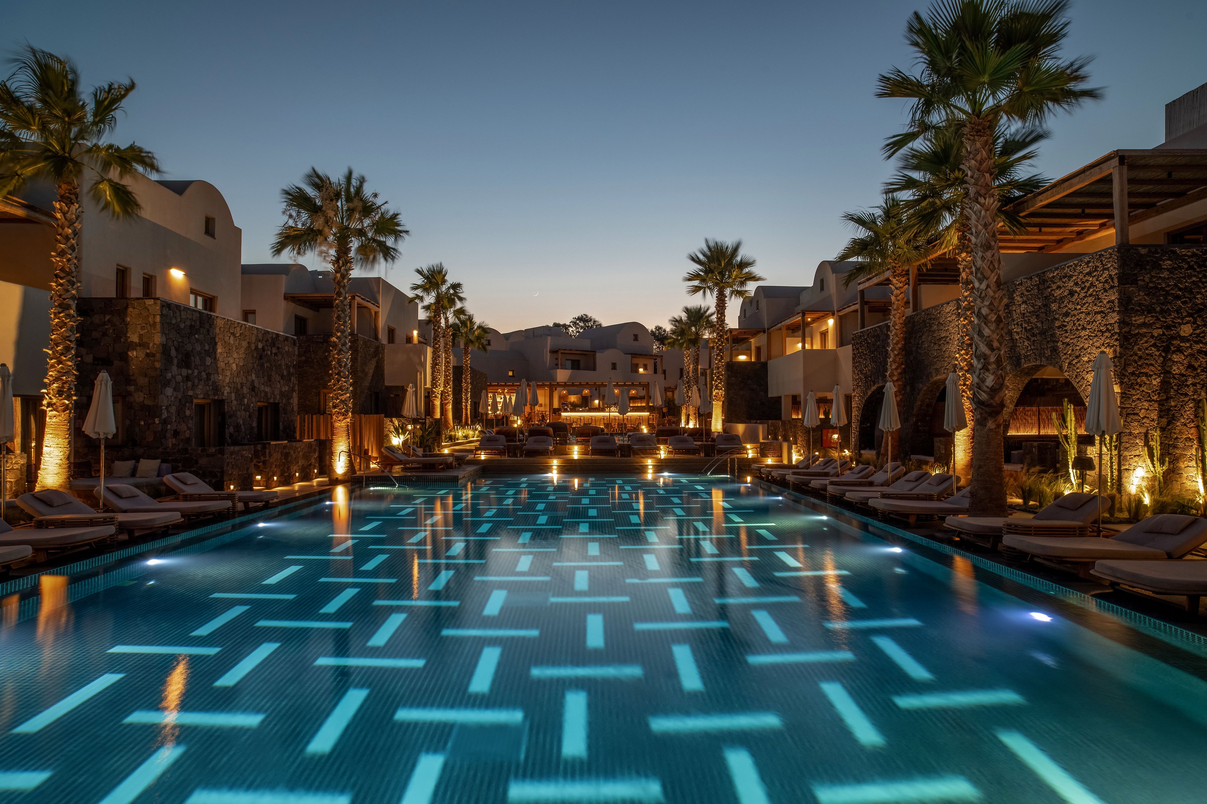 A large resort swimming pool glows with geometric underwater lights at twilight. Palm trees and modern stone buildings line both sides of the pool, creating a symmetrical, inviting scene. Lounge chairs and umbrellas are arranged neatly along the deck, suggesting a relaxing vacation atmosphere. Warm architectural lighting contrasts with the cool blue tones of the water under a clear evening sky.