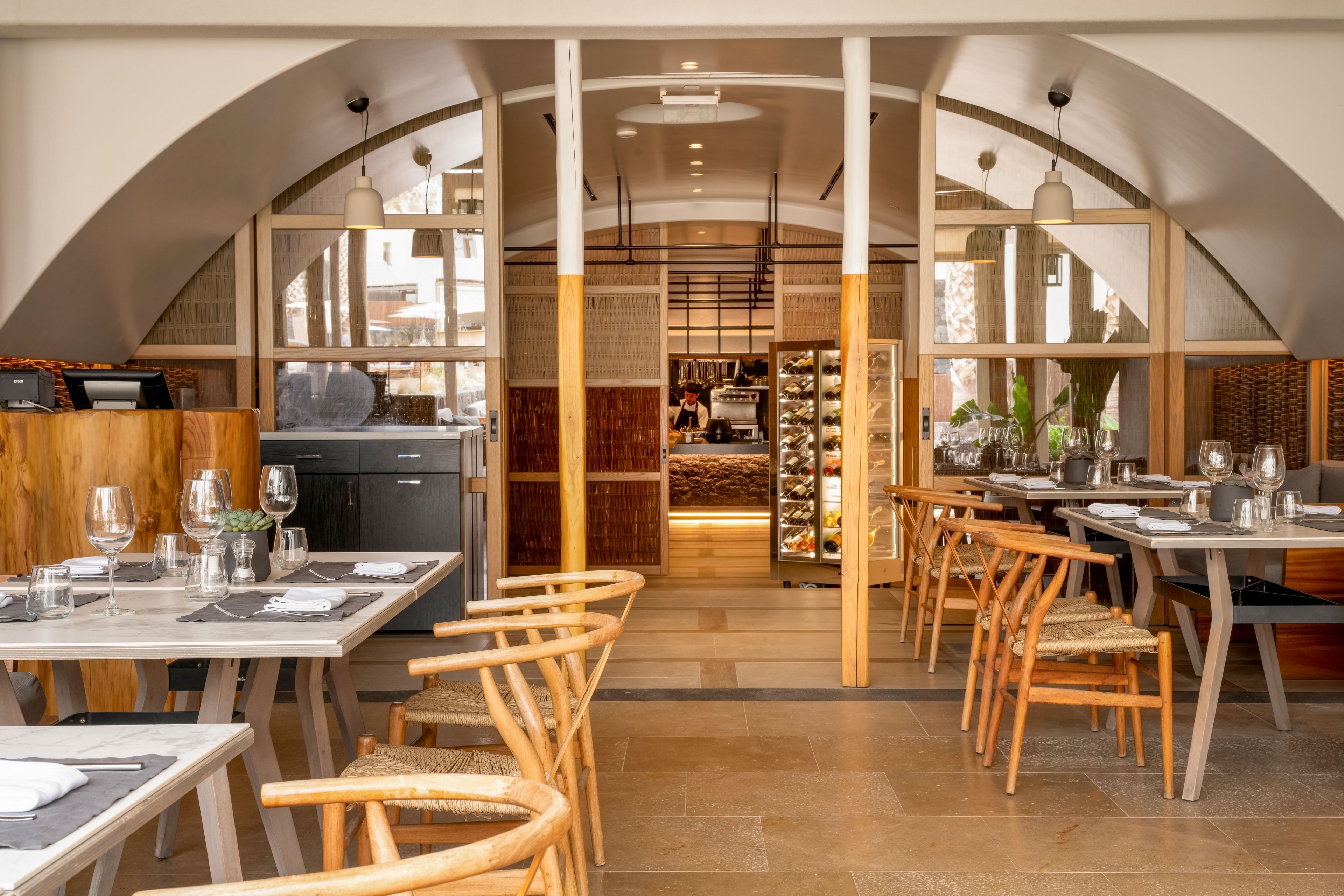 Contemporary restaurant dining area featuring neatly arranged tables and wooden chairs. Place settings with glassware, cutlery, and napkins are prepared for guests, suggesting a fine dining experience. Warm neutral tones, natural wood surfaces, and soft lighting create an inviting atmosphere. In the background, an open kitchen or service area with shelves and a wine or beverage display adds depth to the space.