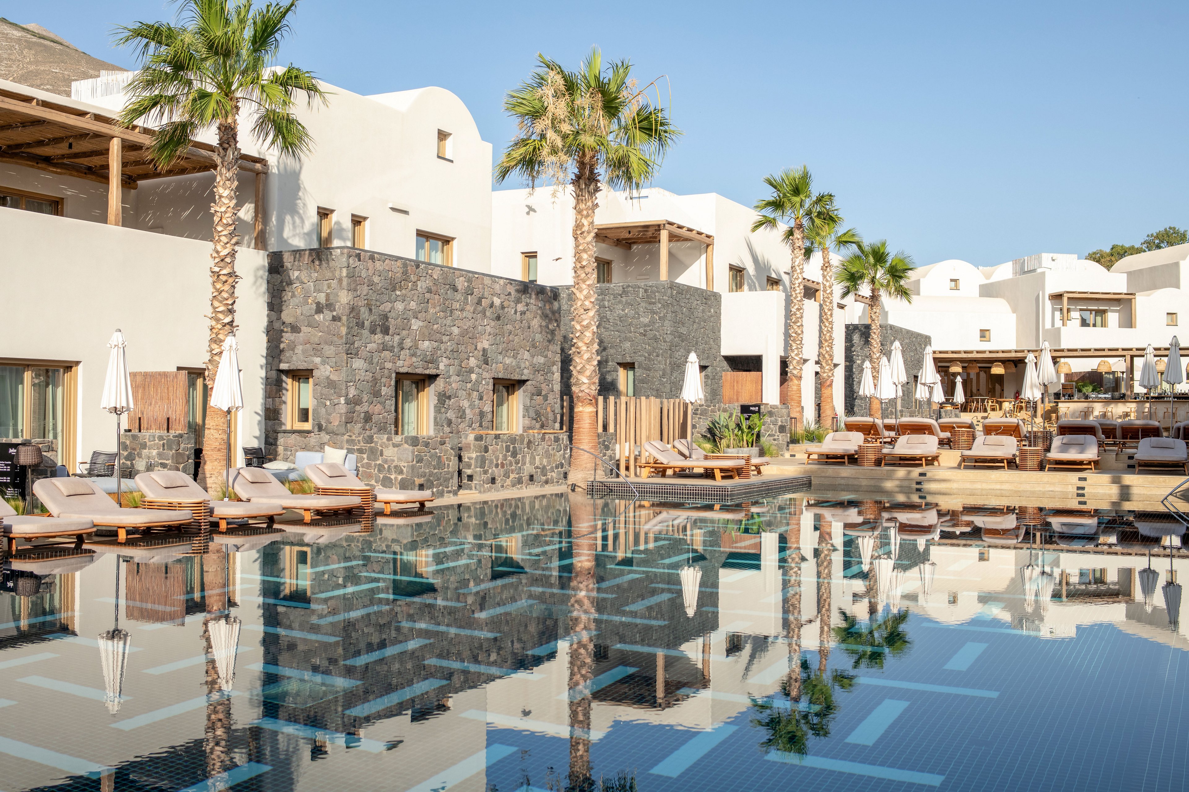 A tranquil resort pool reflects modern white buildings and tall palm trees in the soft morning light. Sun loungers and umbrellas line the poolside, suggesting a relaxing vacation setting. The architecture combines smooth white walls with dark stone accents, creating a contemporary Mediterranean feel. Clear blue water and bright skies enhance the calm, inviting atmosphere of the scene.